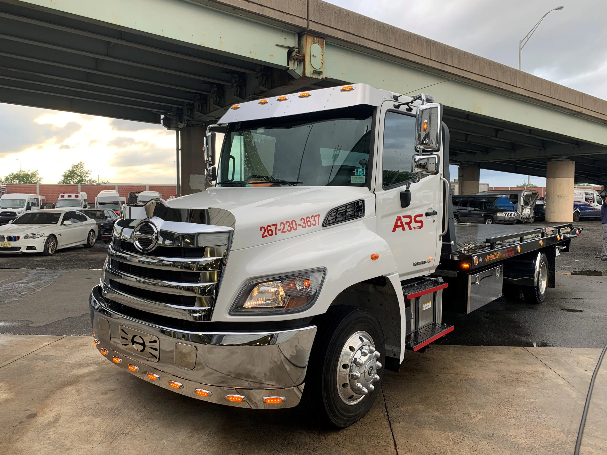 White flatbed tow truck parked under an overpass with other vehicles in the background, cloudy sky overhead.