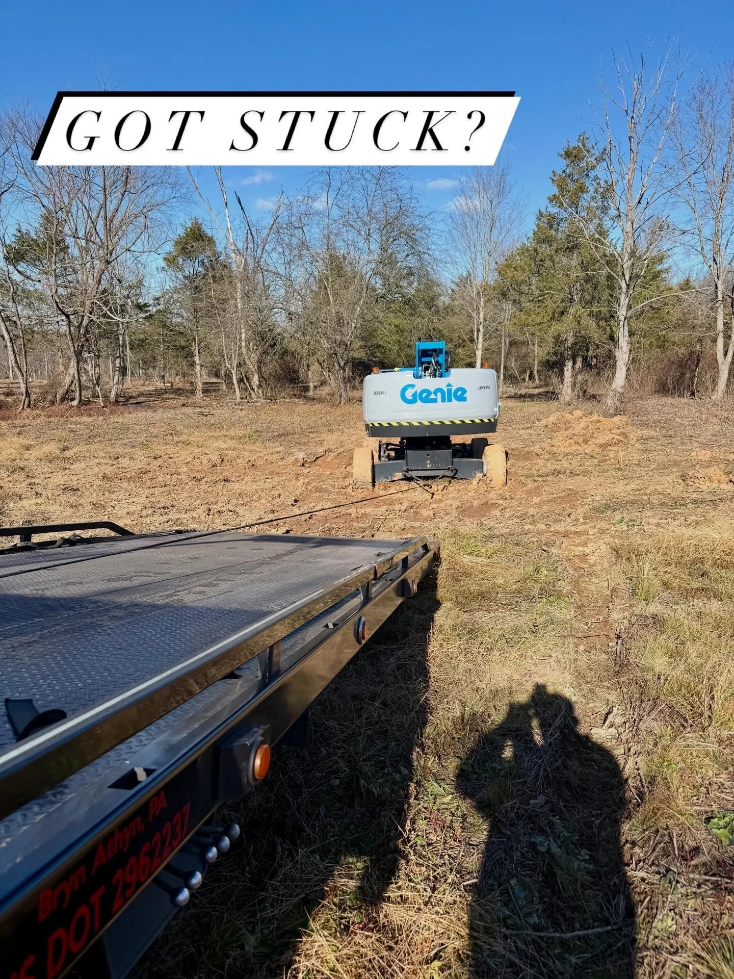 A small Genie excavator in a cleared field with a blue sky overhead. In the foreground, part of a flatbed trailer is visible. Shadows of two people taking the photo are cast on the ground.