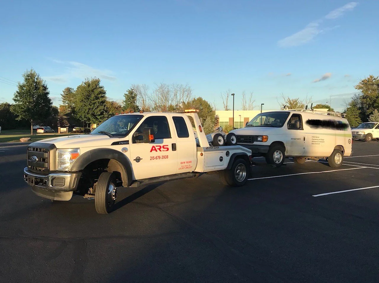 A flatbed tow truck with the logo 'ARS' is towing a white van in a parking lot during daytime.