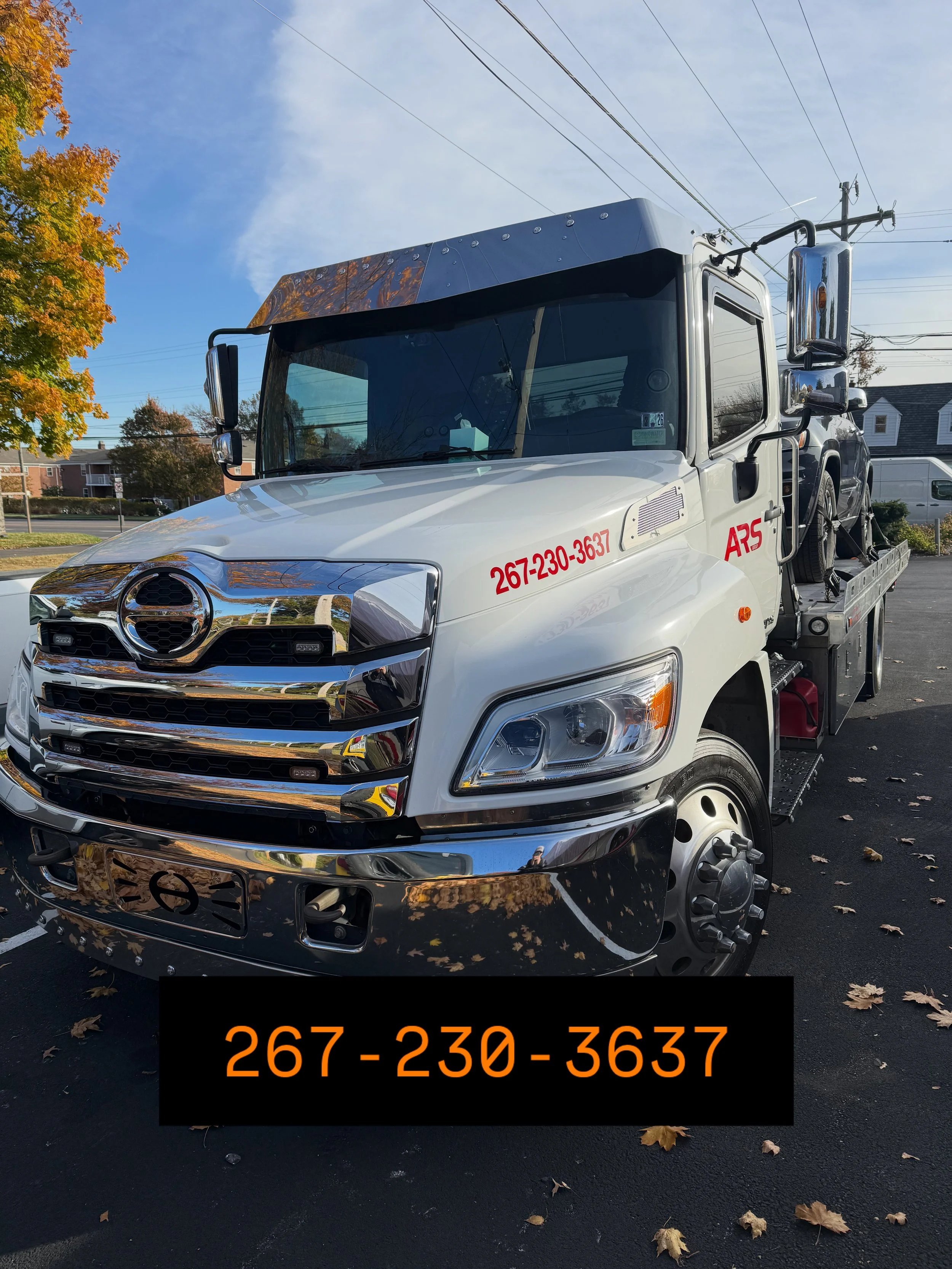 Tow truck parked outdoors with a white cab, black and chrome front grille, and contact information in red text on the side and black background. There are trees with fall leaves and houses in the background.