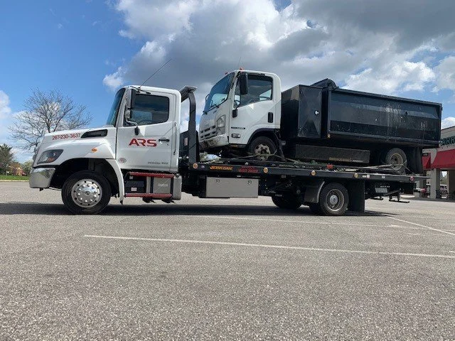 A flatbed tow truck parked in an empty parking lot carrying a small white commercial truck with a black cargo bed.