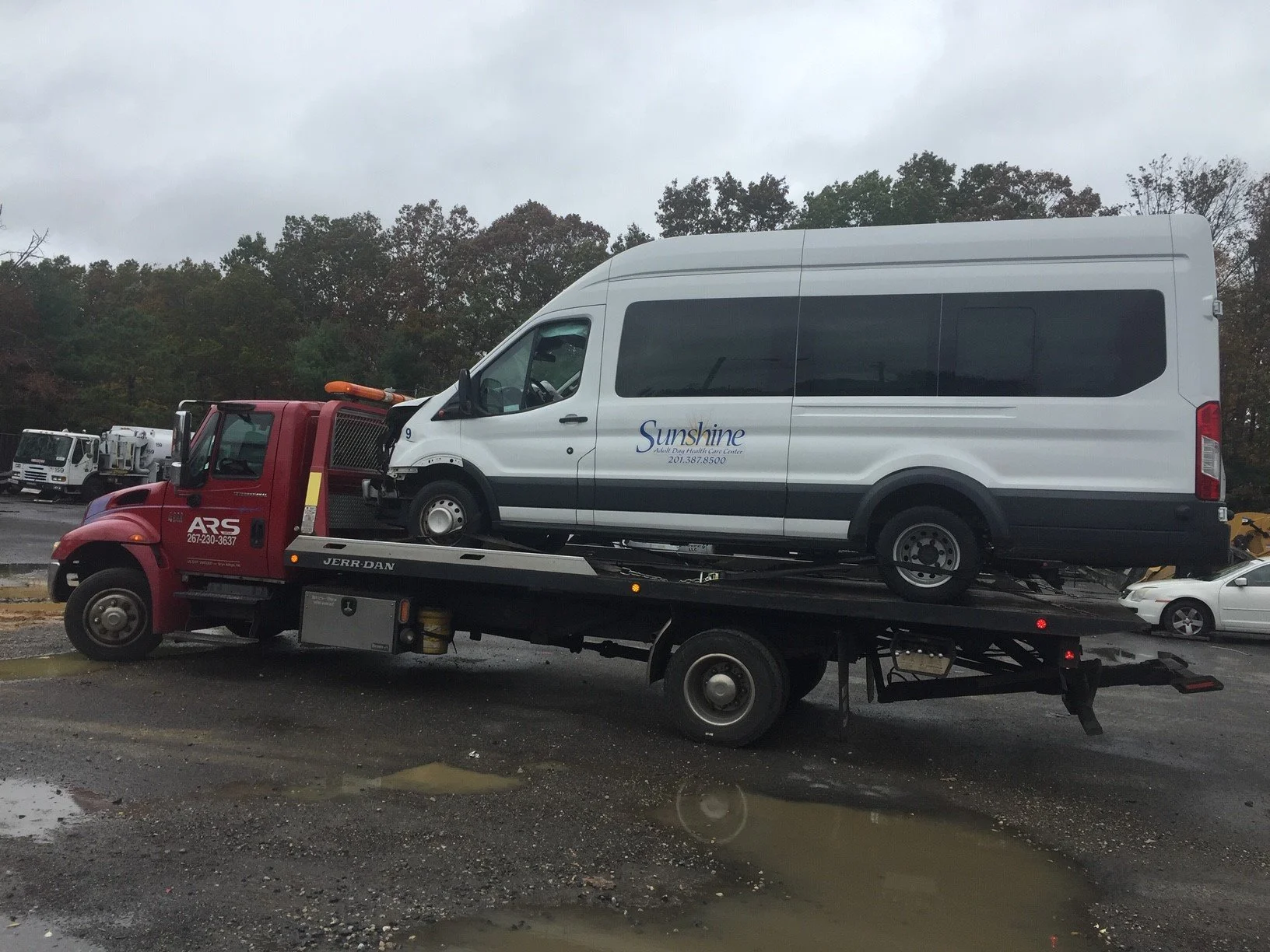 A white van being transported on a flatbed tow truck with a red cab, parked in a lot on a rainy day, with puddles on the ground and trees in the background.
