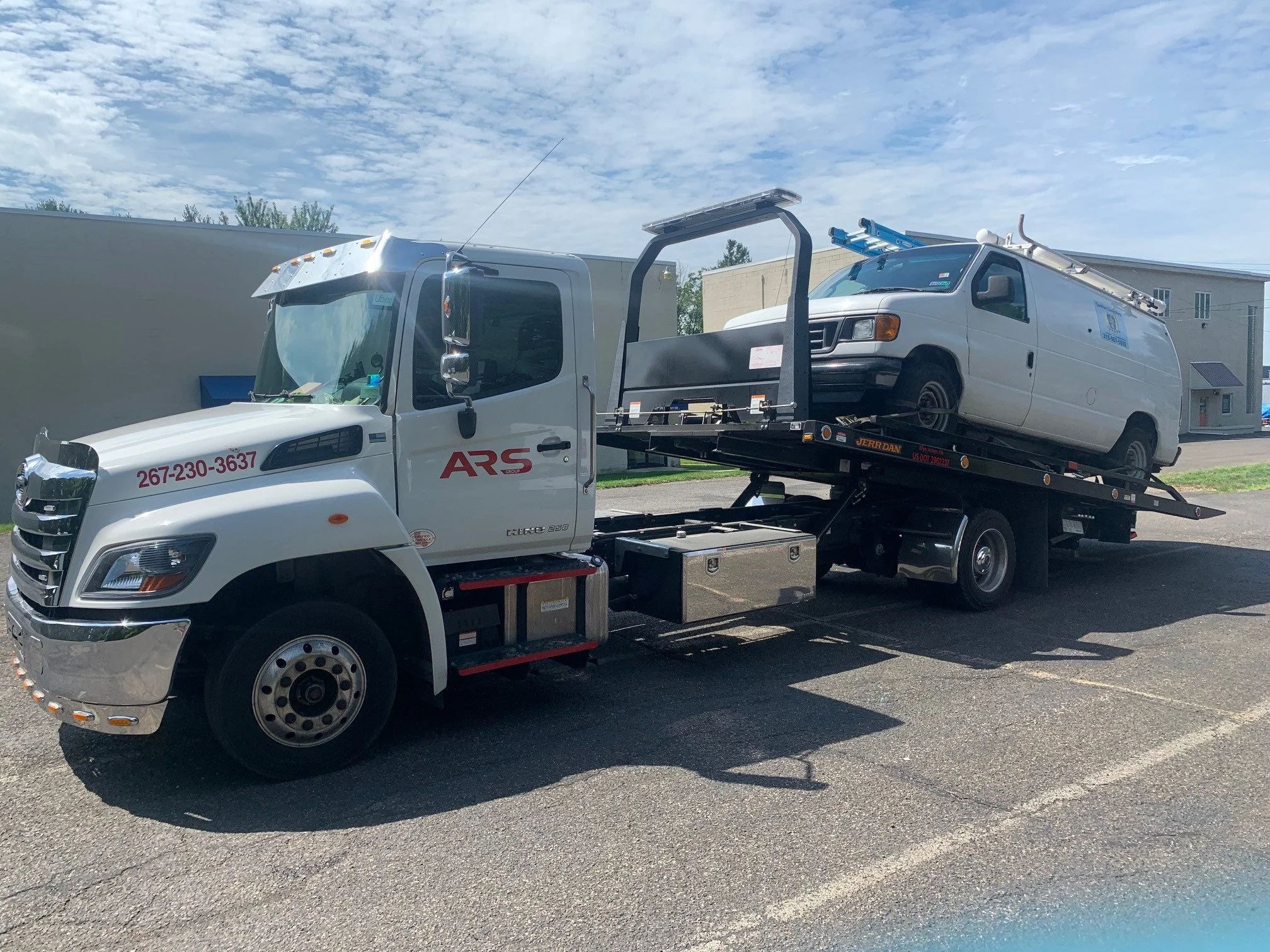 A white tow truck with ARS written on the side is transporting a white utility van on its flatbed, parked on an asphalt lot with a partly cloudy sky above.