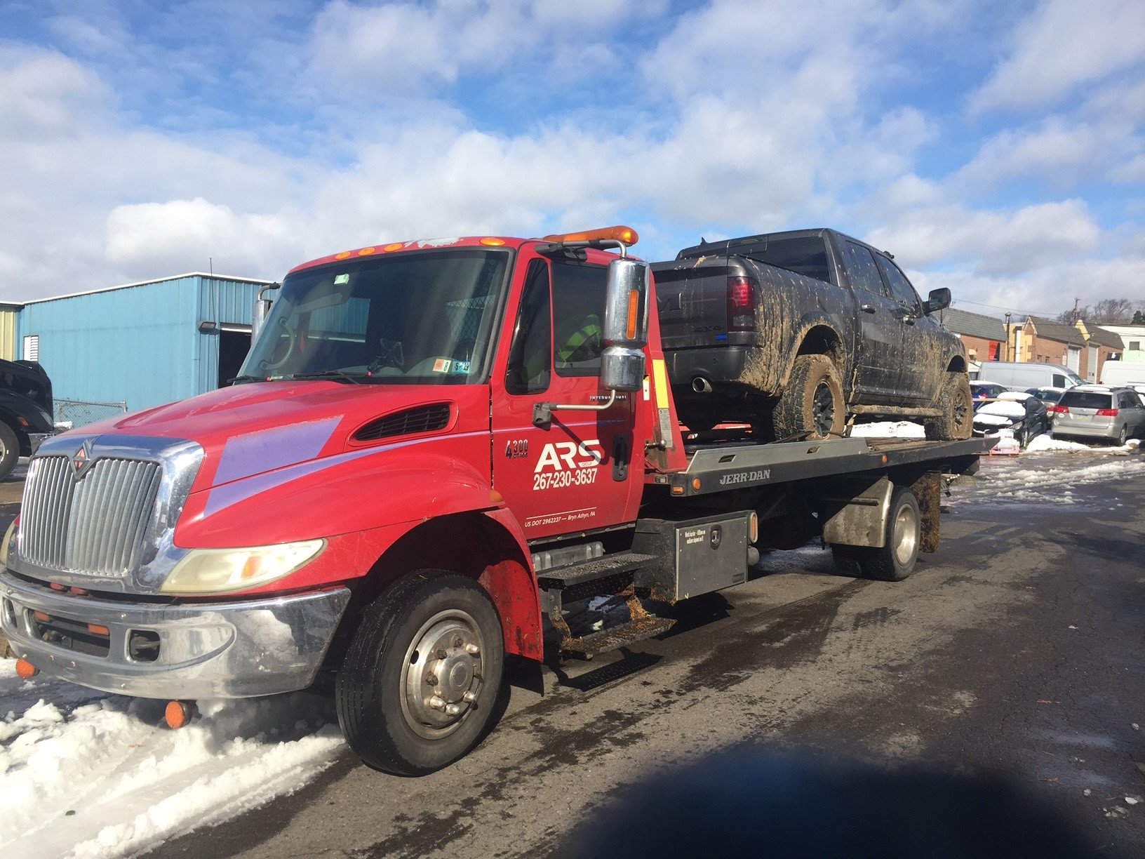 Red tow truck transporting a muddy black pickup truck on a sunny winter day at a parking lot.