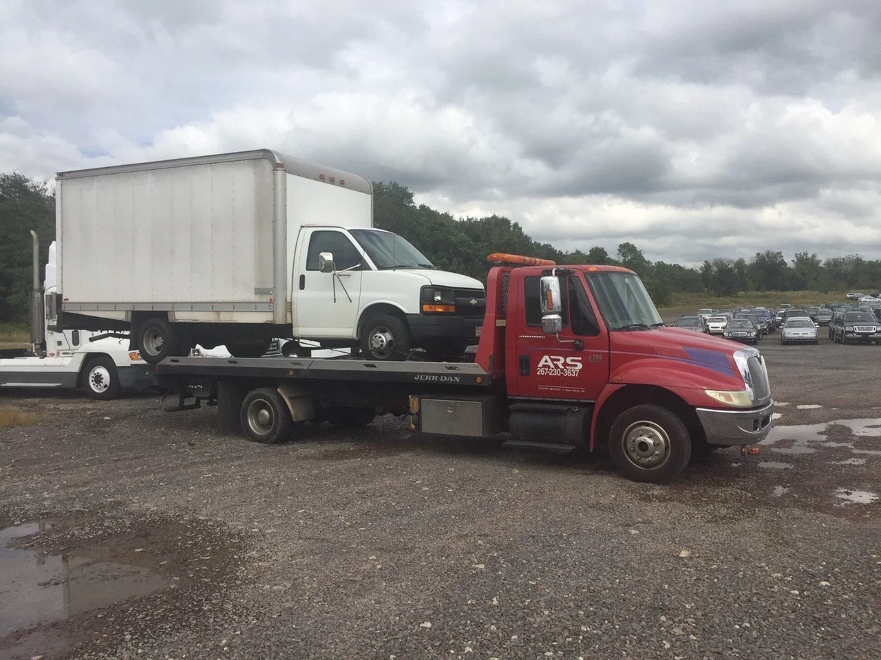A red tow truck transporting a white box truck on a flatbed in a parking lot with many cars, cloudy sky, gravel ground, and trees in the background.