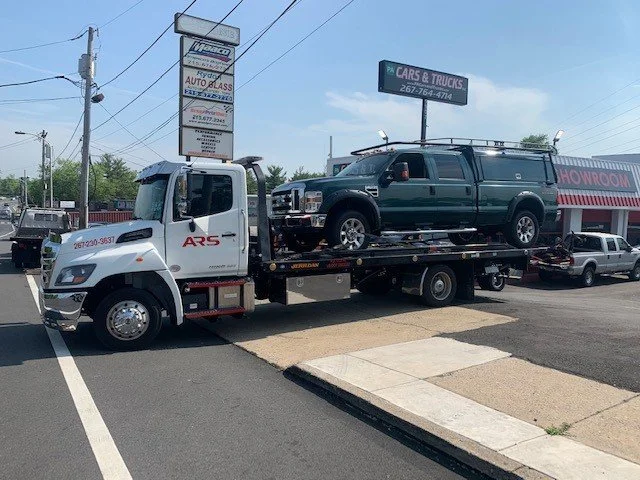 A flatbed tow truck is carrying a black pickup truck with a ladder rack, parked on a street in front of auto service businesses and signage, including a sign for cars and trucks repair.
