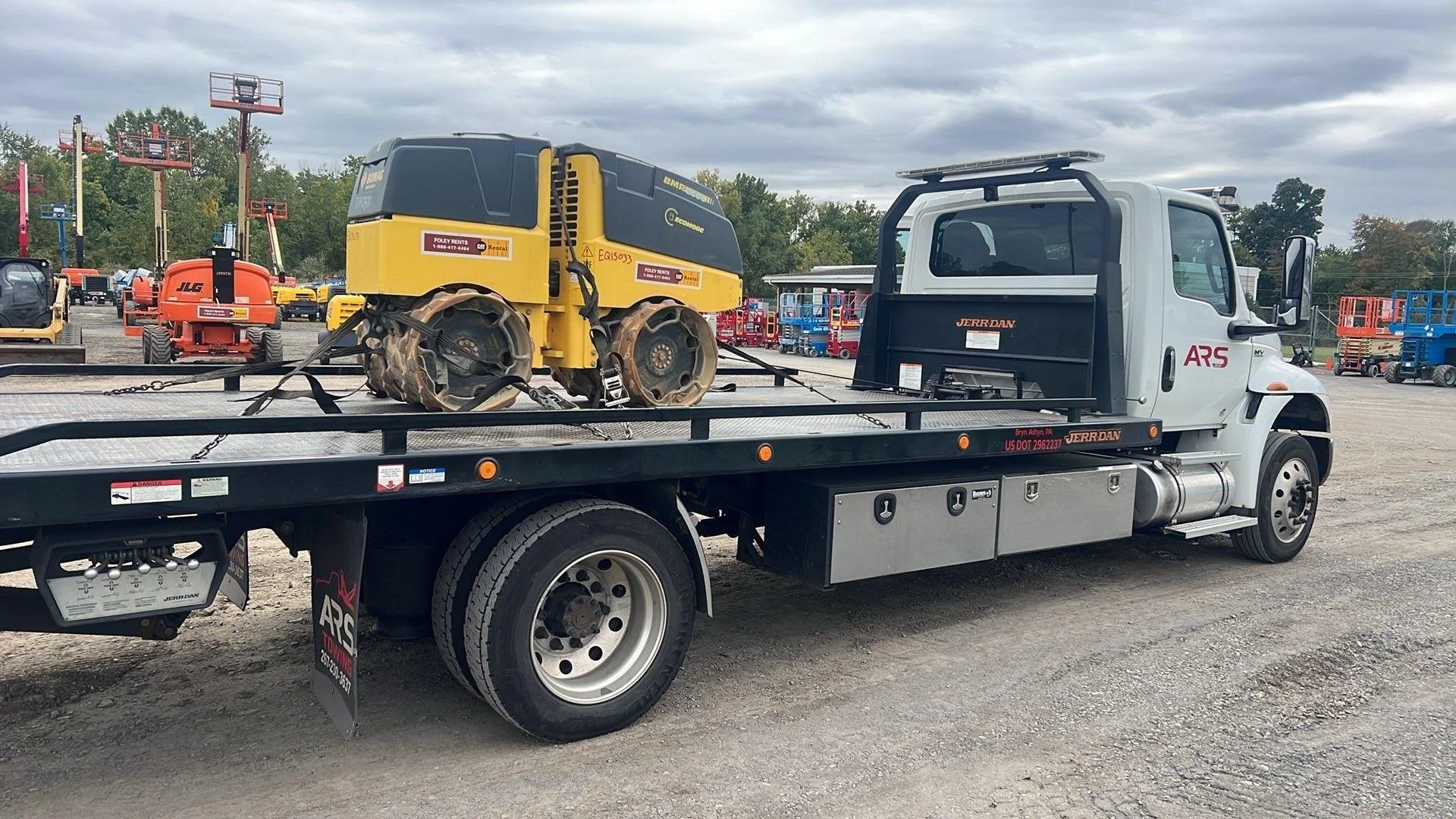 A flatbed tow truck parked in a lot, carrying a small, yellow, remote-controlled construction vehicle with tracks, surrounded by various construction equipment and machinery.