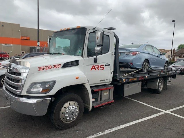 White tow truck with 'ARS' logo carrying a gray sedan in a parking lot, overcast sky.