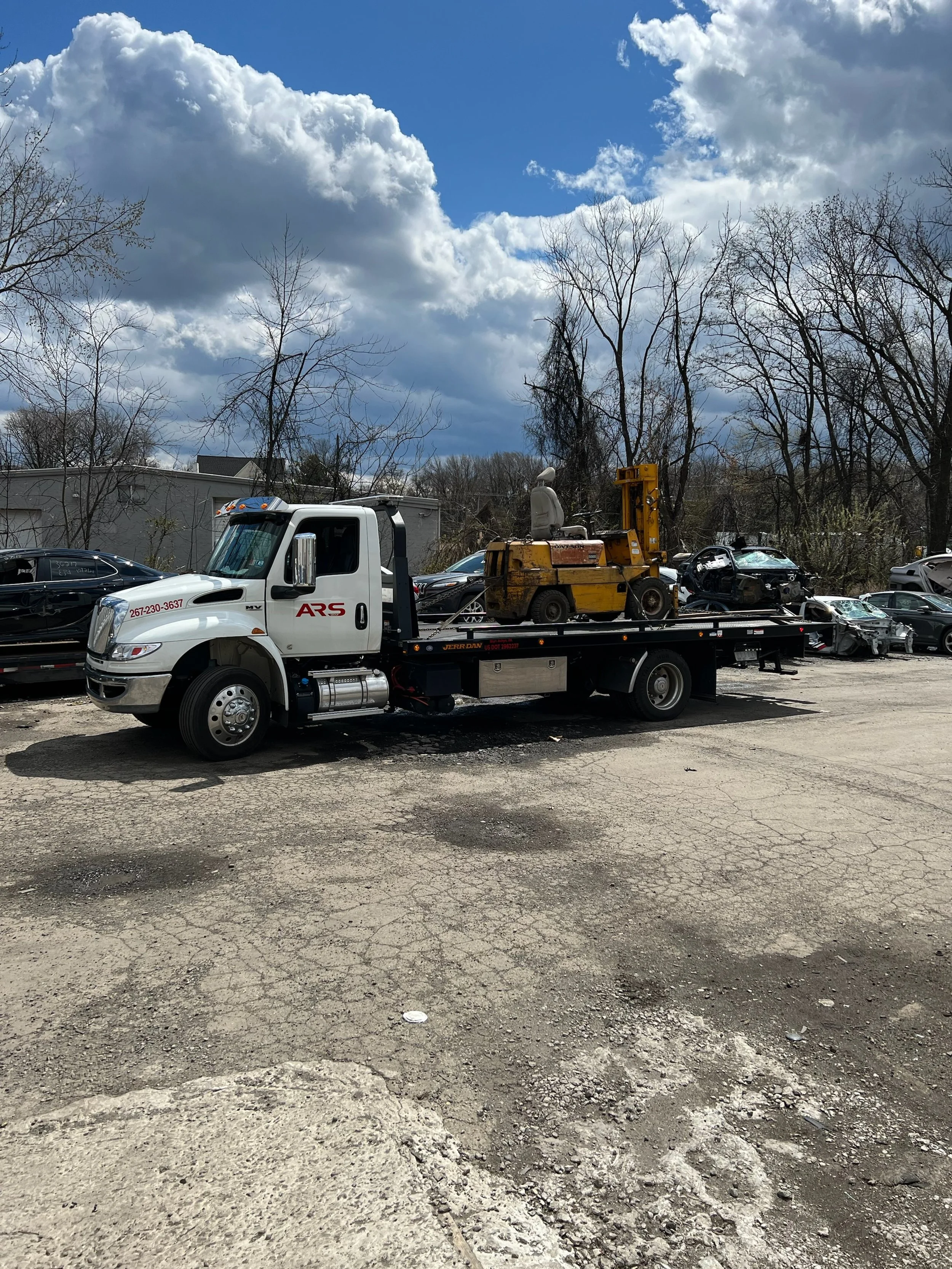 A flatbed tow truck parked in a lot with damaged cars behind it, and a cloudy sky overhead.