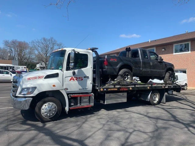 A tow truck carrying a black pickup truck on its flatbed, parked outside in a residential area with houses and trees in the background.