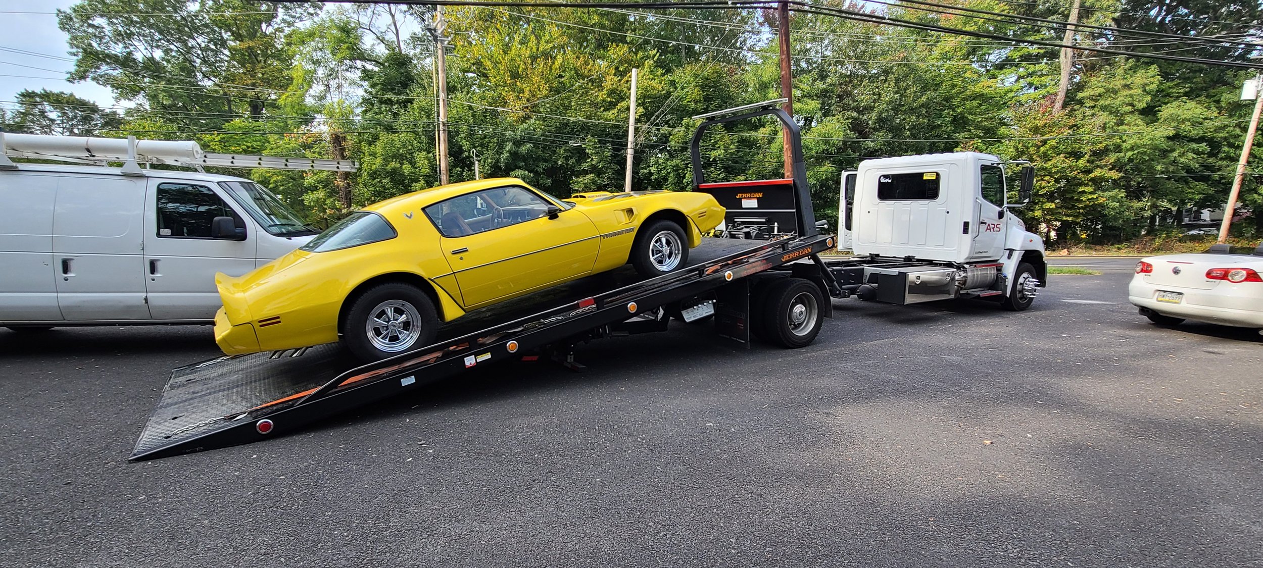 A yellow vintage sports car is being transported on a flatbed tow truck, with a white service van and cars parked nearby, and trees in the background.