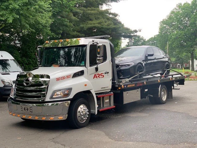 A tow truck transporting a black sedan on a flatbed, parked on a suburban street with trees in the background.