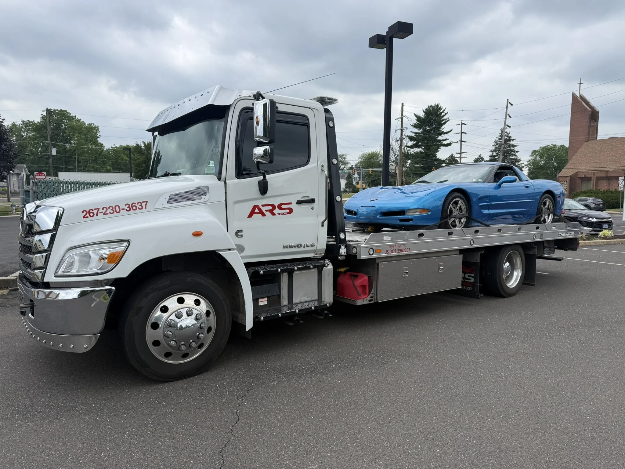 ARS Towing white flatbed tow truck transporting a blue convertible sports car from a parking lot.
