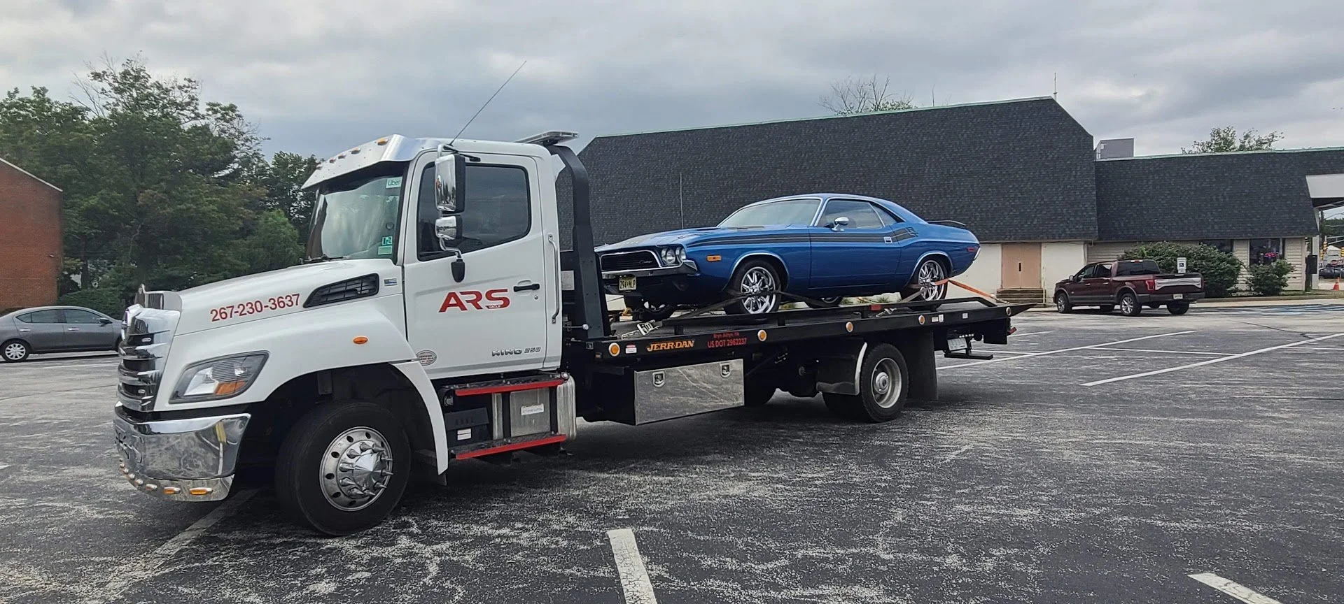 A white tow truck carrying a blue vintage muscle car in an empty parking lot with a building and trees in the background.
