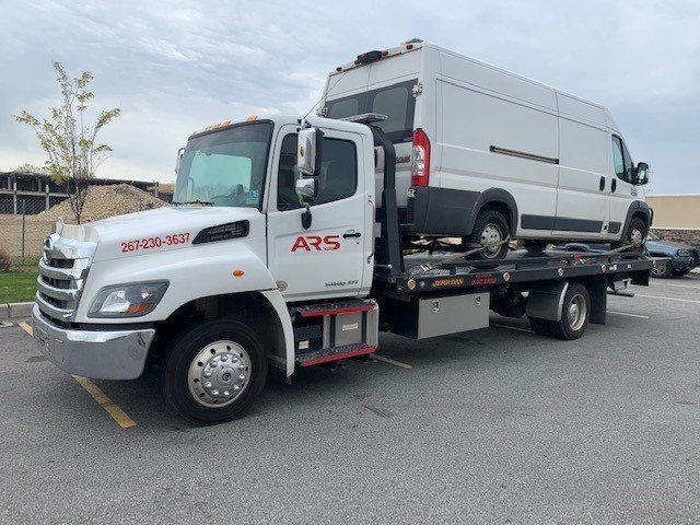 A white tow truck transporting a white cargo van on its flatbed in a parking lot.
