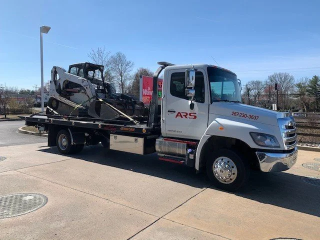 A tow truck carrying a skid-steer loader on its flatbed in a parking lot.
