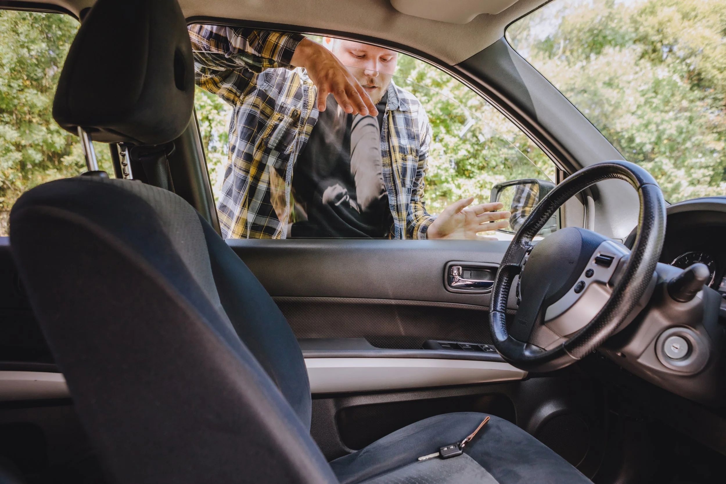 A man standing outside a car talking to a woman inside, seen from the car's back seat, with trees in the background.