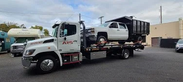 A tow truck transporting a white pickup truck with a black bed in a parking lot.