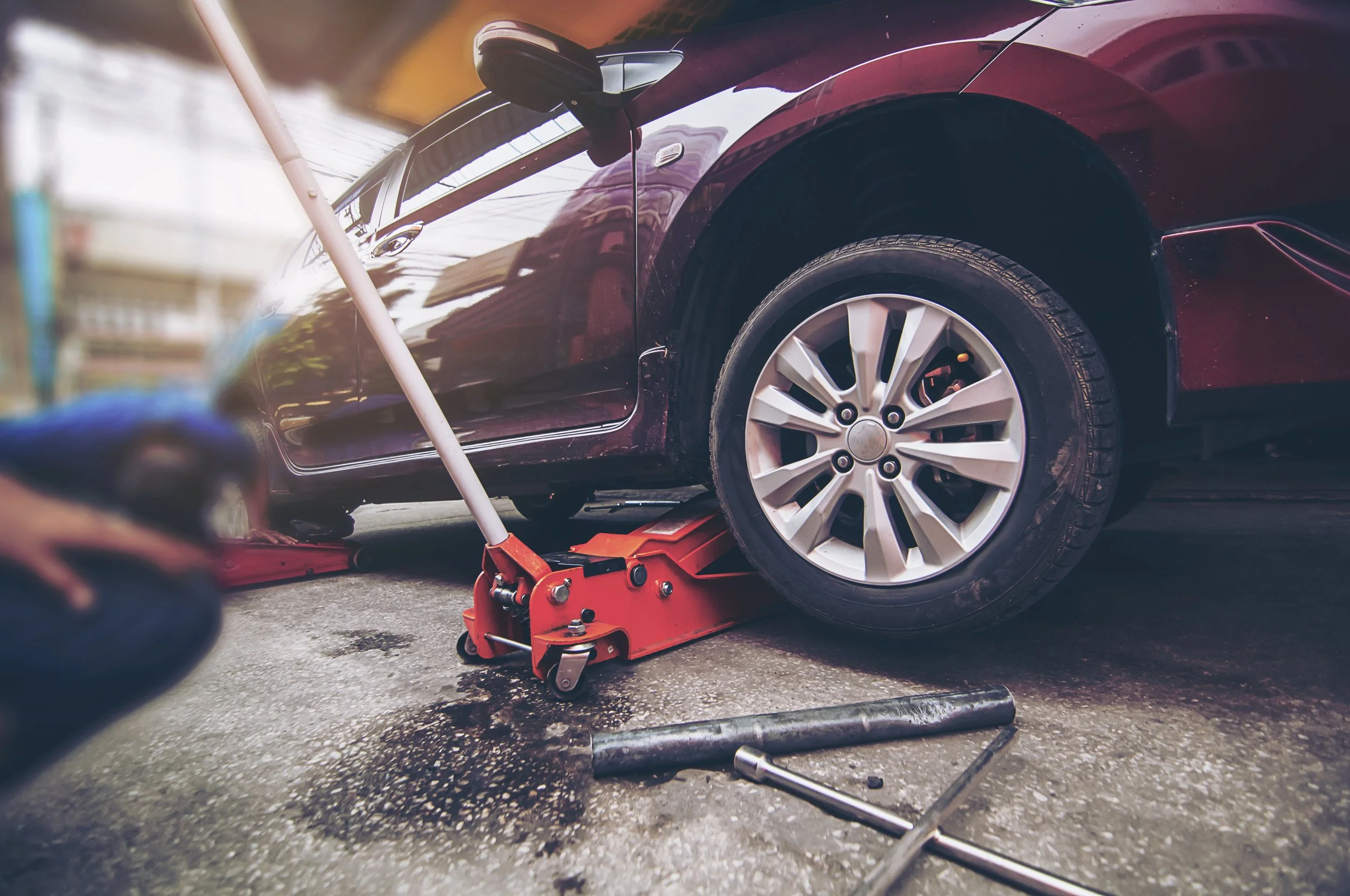 A person changing a flat tire on a maroon car using a hydraulic jack and a lug wrench on a paved surface.