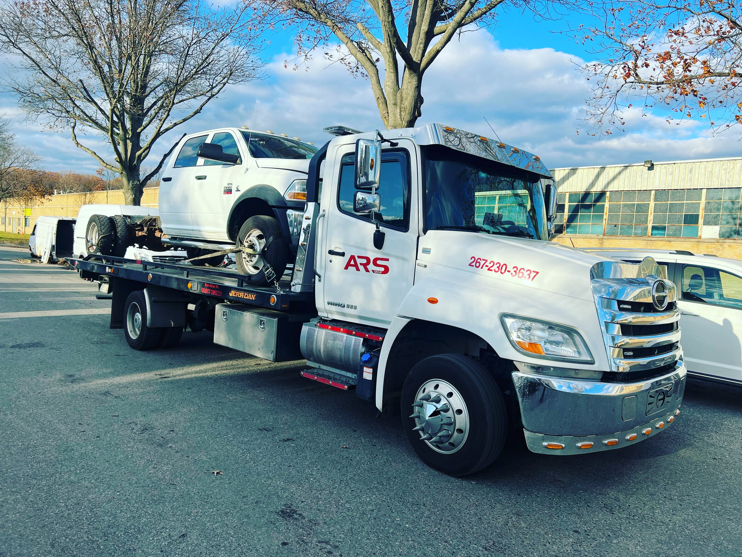 A white tow truck transporting a white pickup truck on a flatbed in a parking lot with trees and a building in the background.
