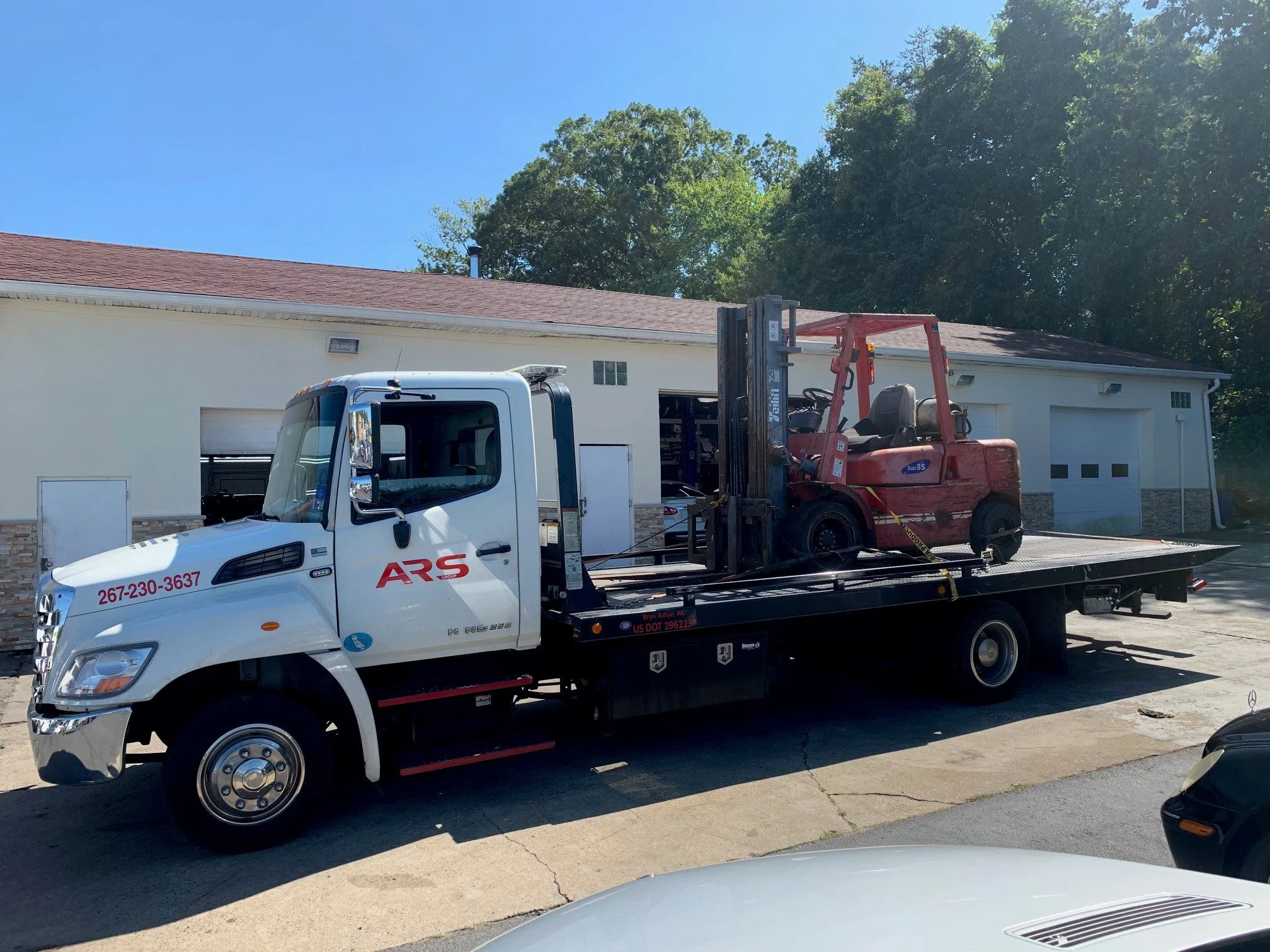 Flatbed tow truck carrying a red forklift outdoors in front of a white building with garage doors under a clear sky.