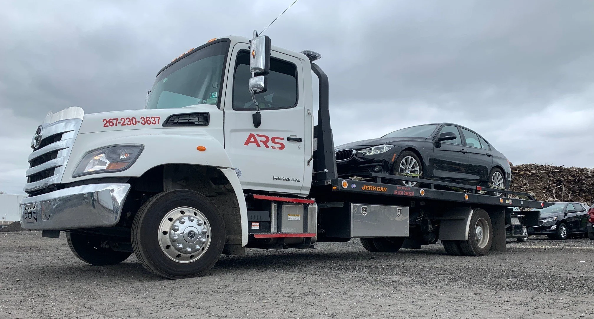 A flatbed tow truck transporting a black sedan in a parking lot, with other vehicles and a cloudy sky in the background.
