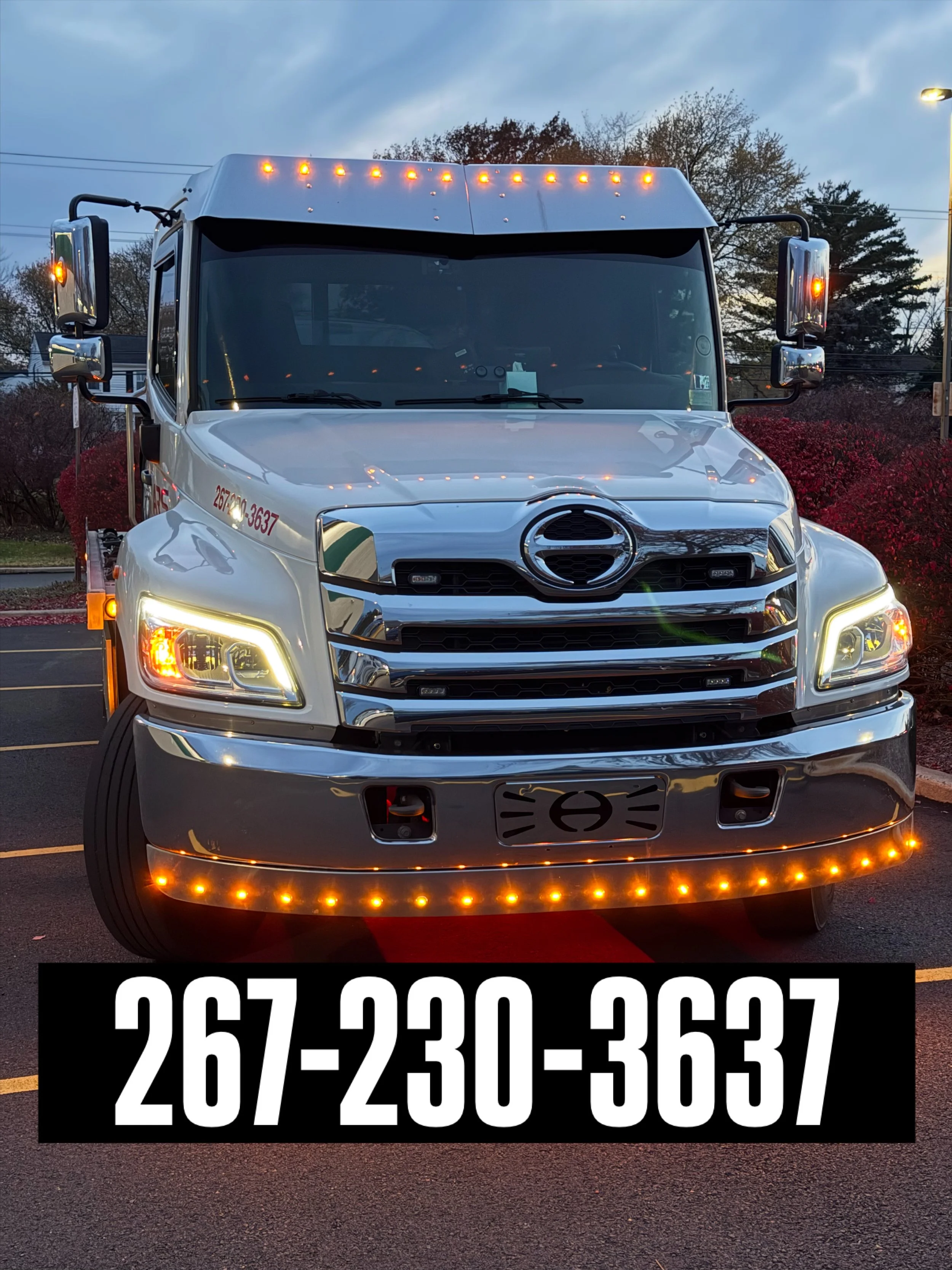 Front view of a white tow truck with amber running lights, large side mirrors, and a black license plate area, parked on a street during twilight, with a red carpet underneath. The truck features a company phone number, 267-230-3637, displayed in white text on a black background at the bottom of the image.