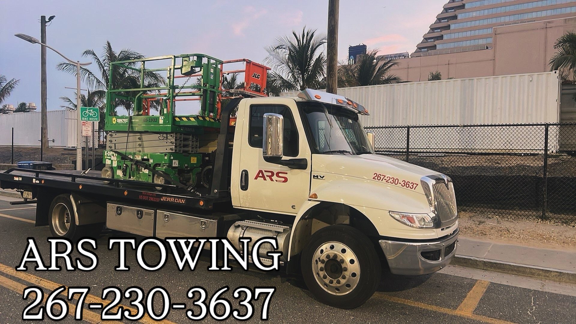 Tow truck labeled 'ARS' parked on street, carrying a green and orange scissor lift. There is a sidewalk, fence, and buildings in the background, with palm trees and a streetlight.