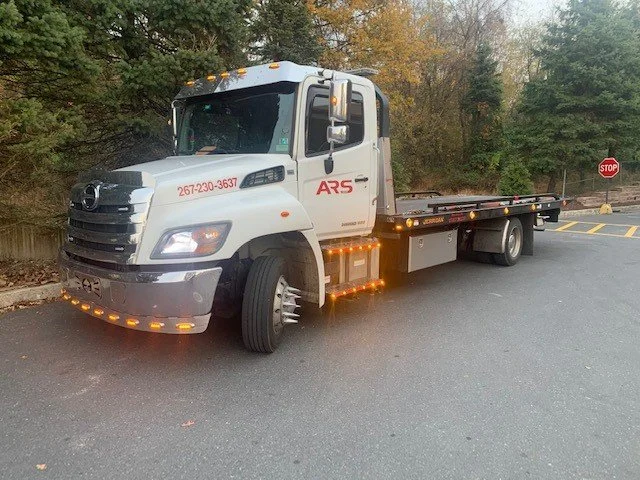A white flatbed tow truck with orange lights parked on a road near trees and a stop sign.
