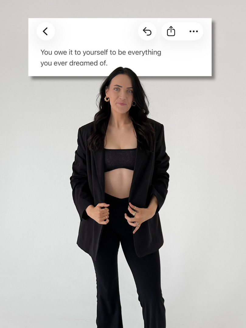 A woman with long dark hair wearing a black blazer, black pants, and a black crop top poses against a plain white wall. There is a quote overlay above her that reads, "You owe it to yourself to be everything you ever dreamed of."
