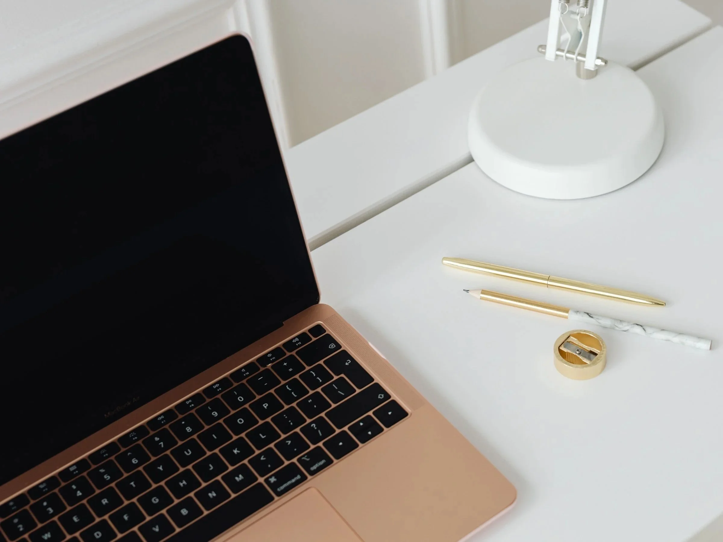 A workspace with a gold-colored laptop, two gold pens, a marble-patterned white pencil, a gold tape measure, and a white desk lamp on a white desk.