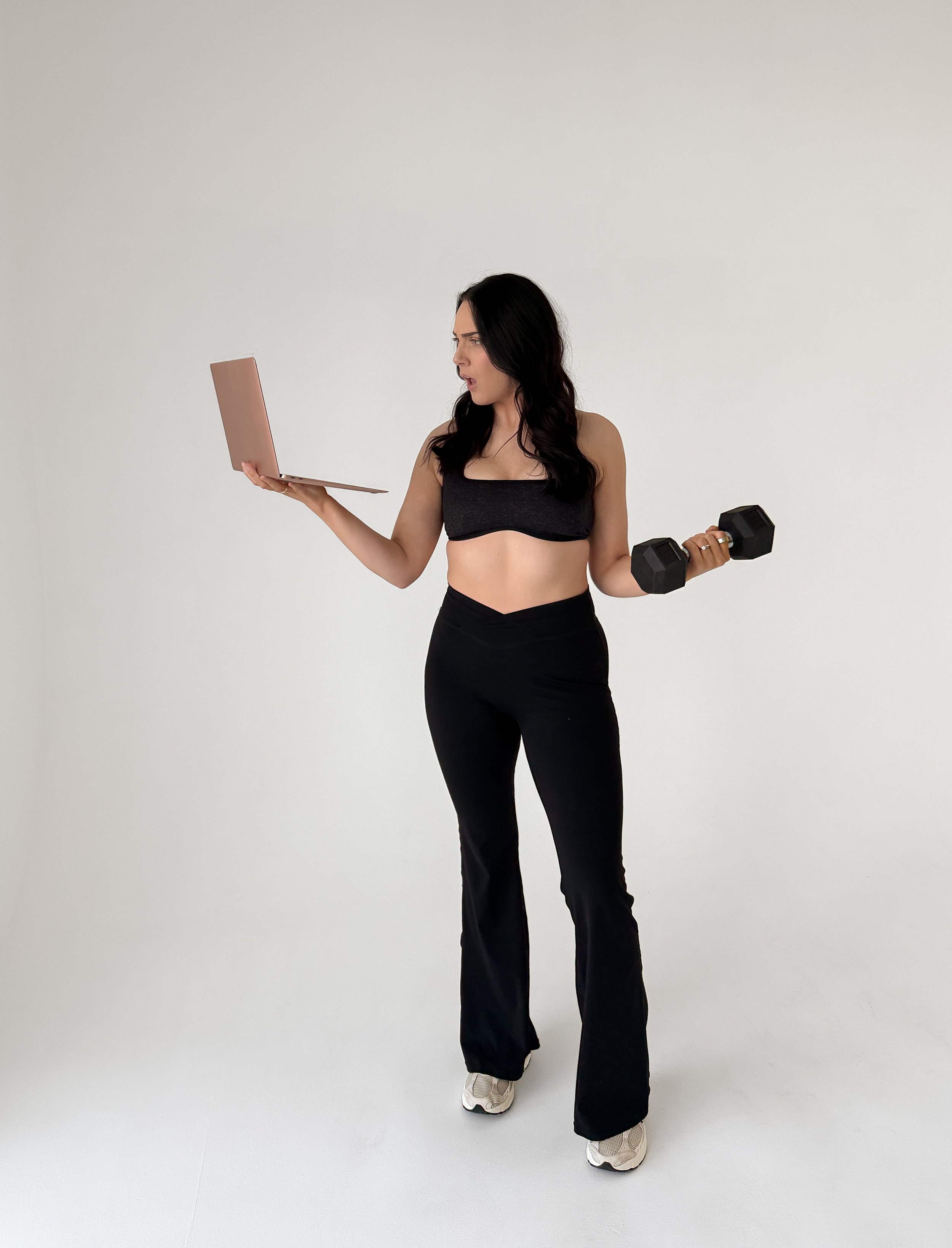A woman holding a dumbbell in one hand and a laptop in the other, standing against a plain white background.