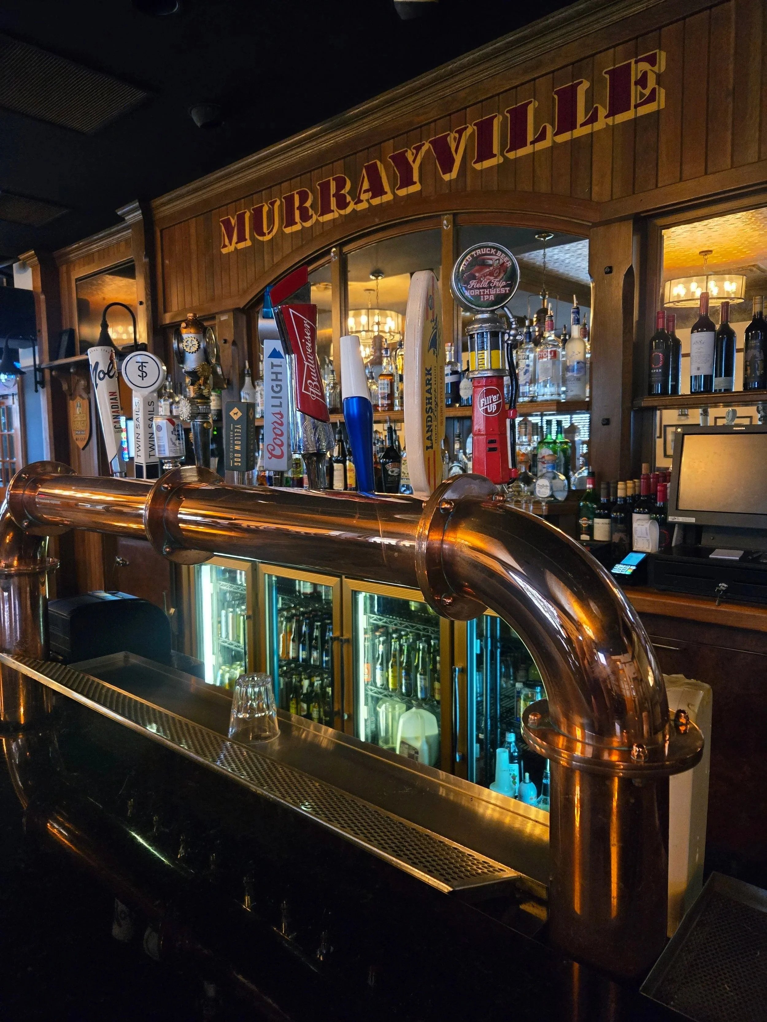 A bar with a wooden arch that has 'MURRAYVILLE' written on it in large red and yellow letters. The bar counter features taps for draft beer, with various bottles of alcohol displayed behind. Copper pipes are part of the bar's draft system, and there is a glass with a beverage or water placed on the counter. The background shows a cozy, warmly lit interior with a chandelier and wooden decor.