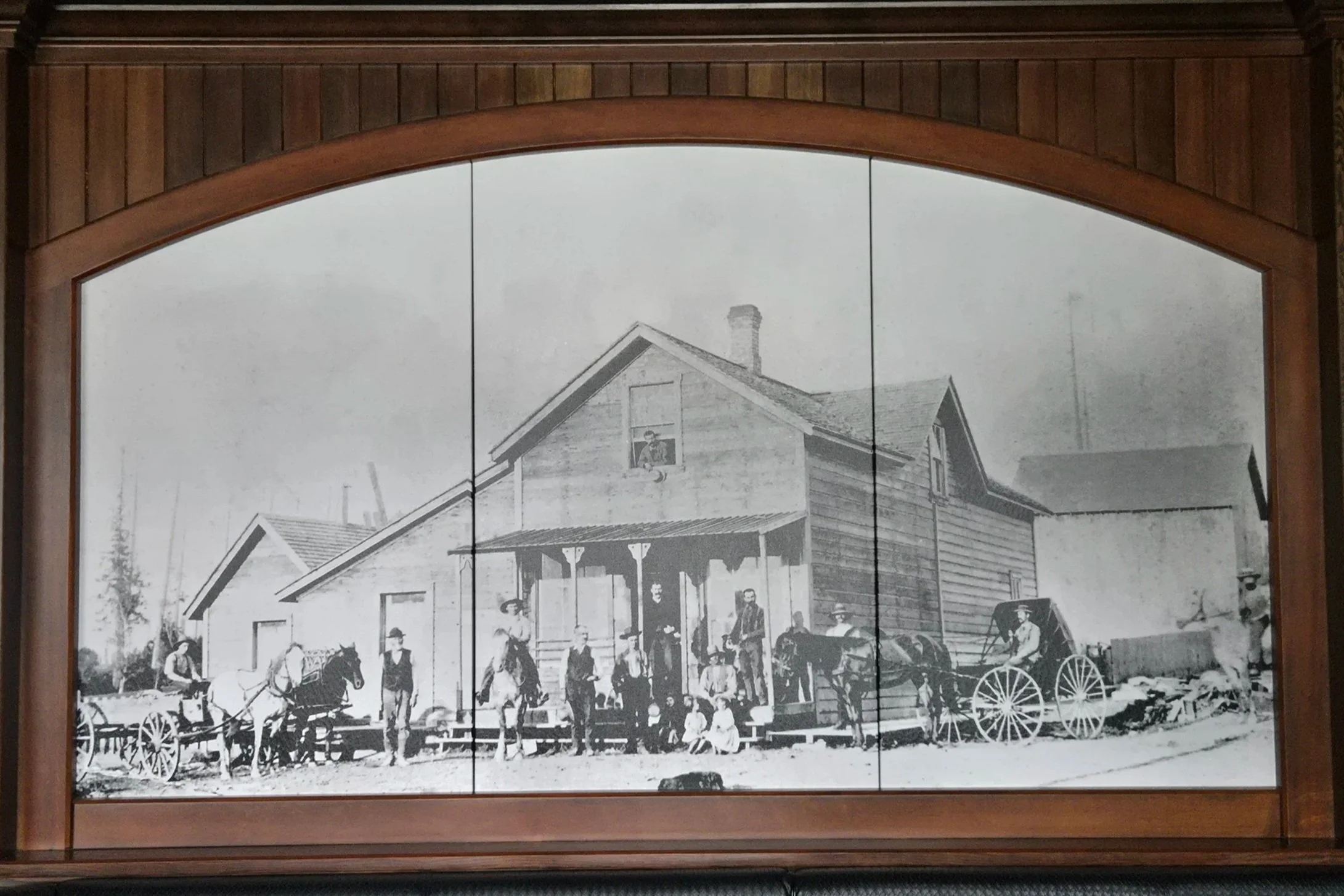Black and white historical photograph of a wooden house with a large porch, surrounded by people, horse-drawn carriages, and wagons, framed by a wooden arch.