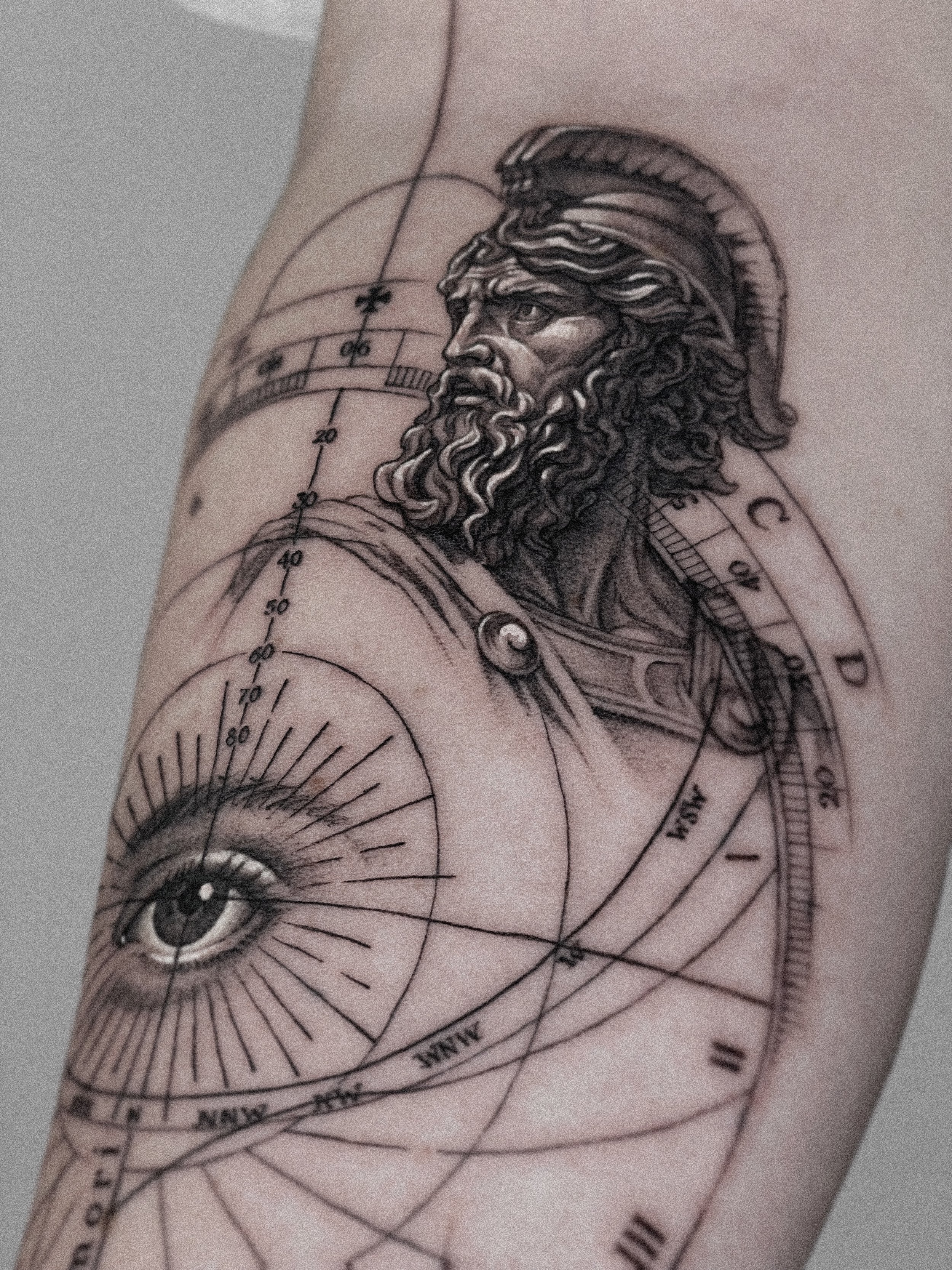 Tattoo featuring a classical statue of a beard and helmeted man, an eye with rays, and technical geometric lines and measurements.