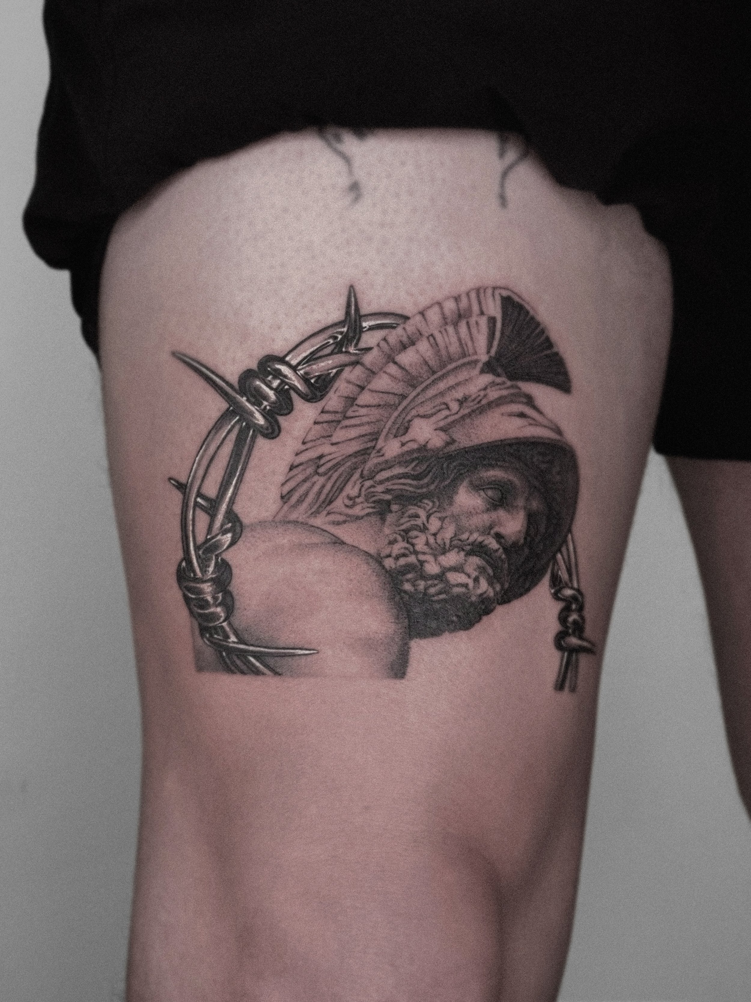 Tattoo of a classical warrior with a helmet and beard, surrounded by barbed wire, on a person's thigh.