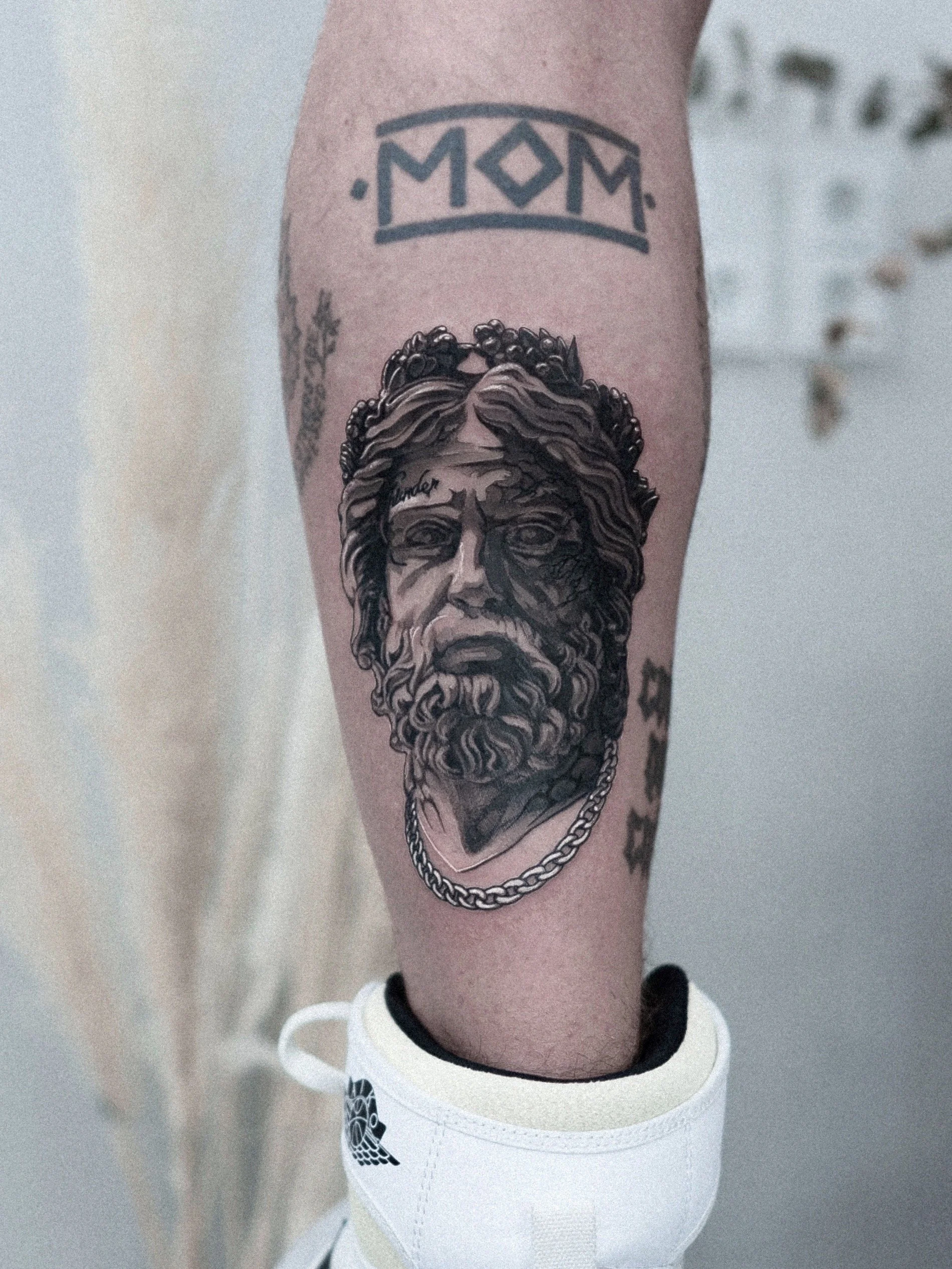 Tattoo of a bearded ancient Greek or Roman statue's face with a chain necklace 