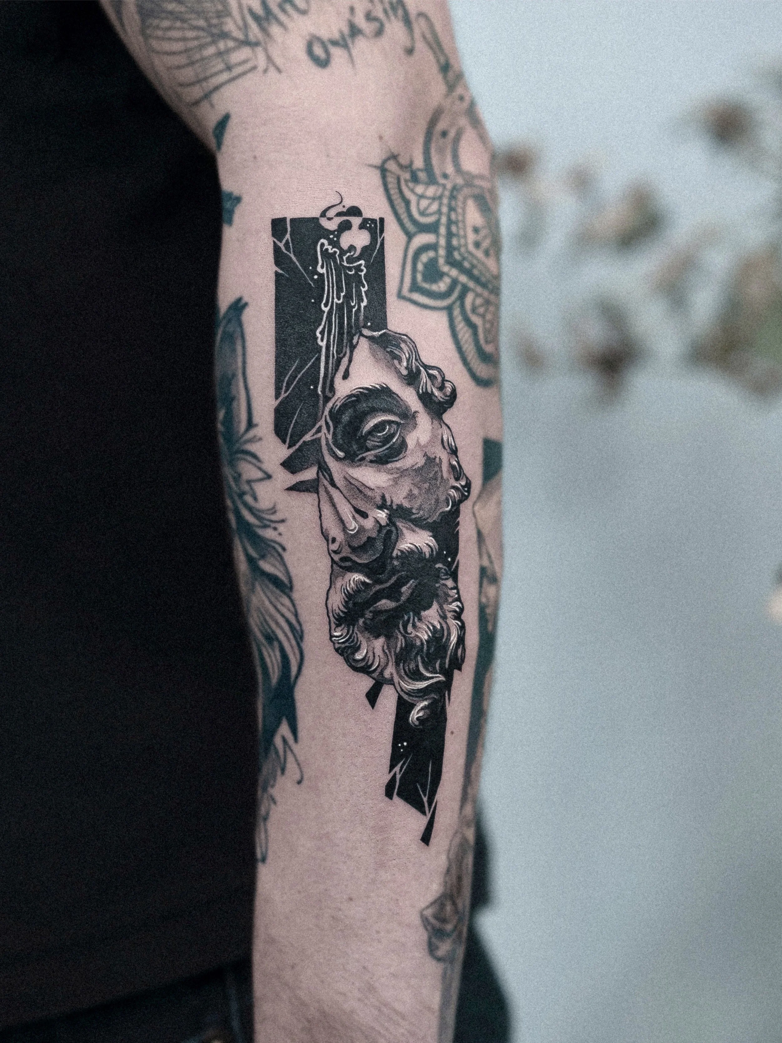 Tattoo of a classical statue's head with a beard, integrated with abstract geometric shapes and lines, on a person's arm.