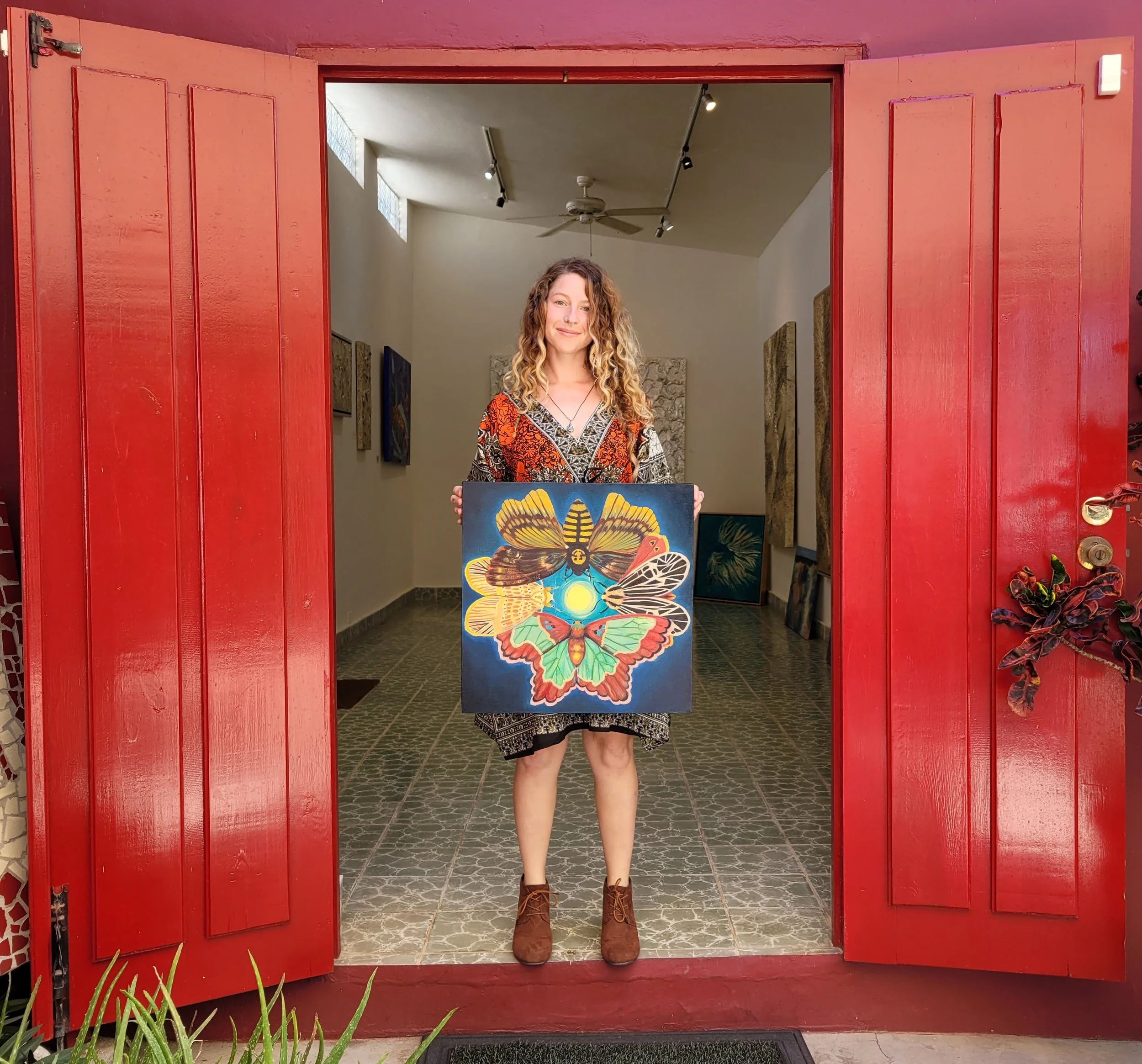 A woman with curly hair, wearing a patterned dress and brown boots, stands in a doorway holding a painting of colourful moths in a circle around the full moon, inside an art gallery with white walls and art pieces displayed on the walls.