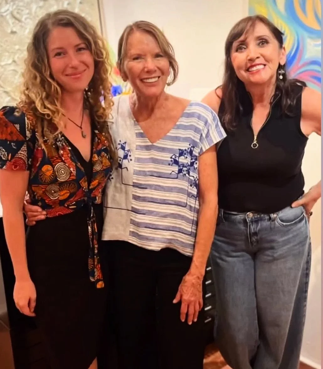 Three women standing together indoors, smiling for a photo. The woman on the left has curly blonde hair and is wearing a colorful patterned top. The woman in the center has short light brown hair and is wearing a striped shirt with blue floral embroidery. The woman on the right has long dark hair and is wearing a black sleeveless top with gray jeans. They are in front of a colorful abstract painting.
