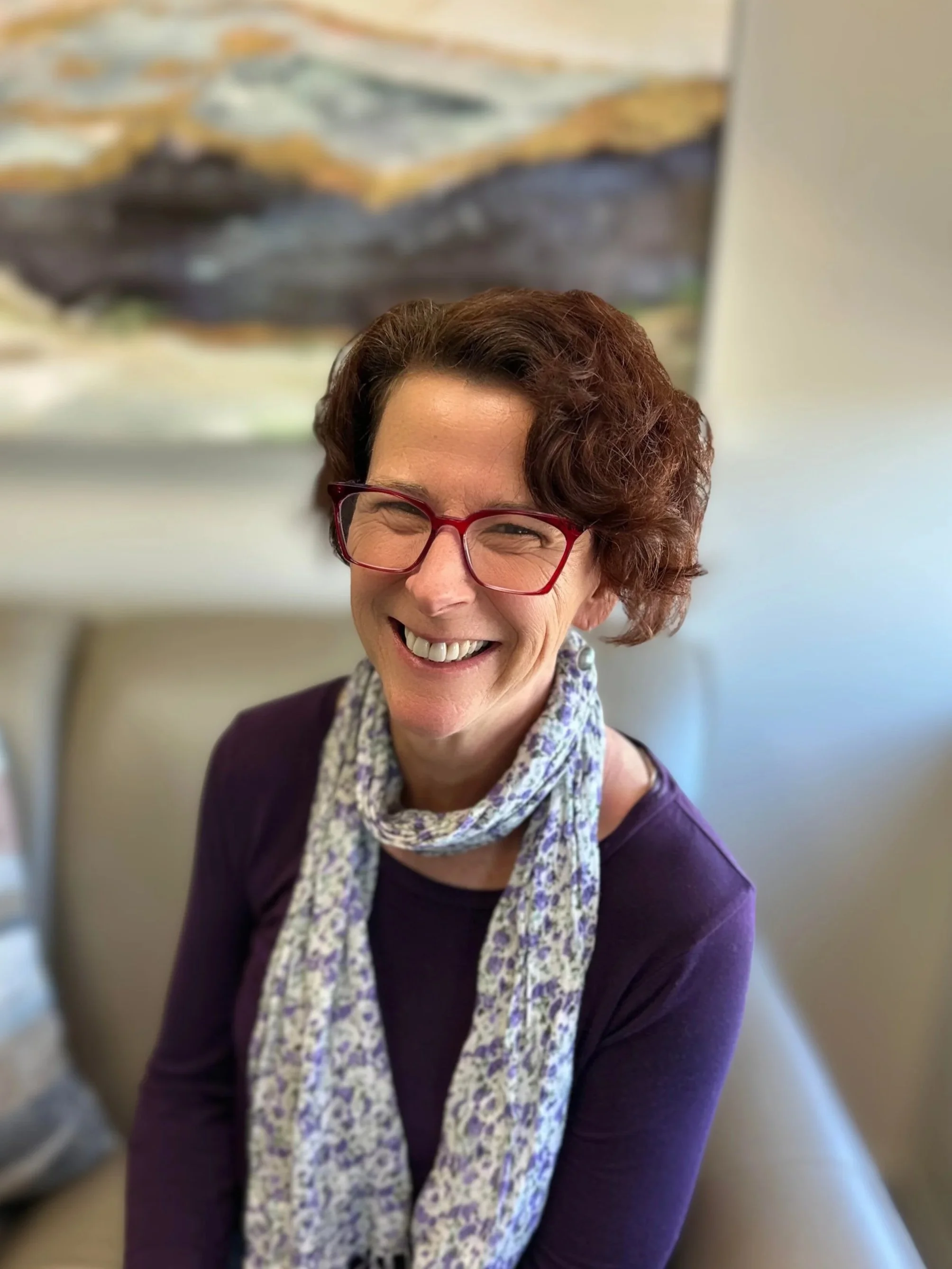 A woman with short curly red hair, wearing red glasses, a purple long-sleeve shirt, and a light patterned scarf, smiling and sitting on a light-colored bench with a blurred landscape painting in the background.
