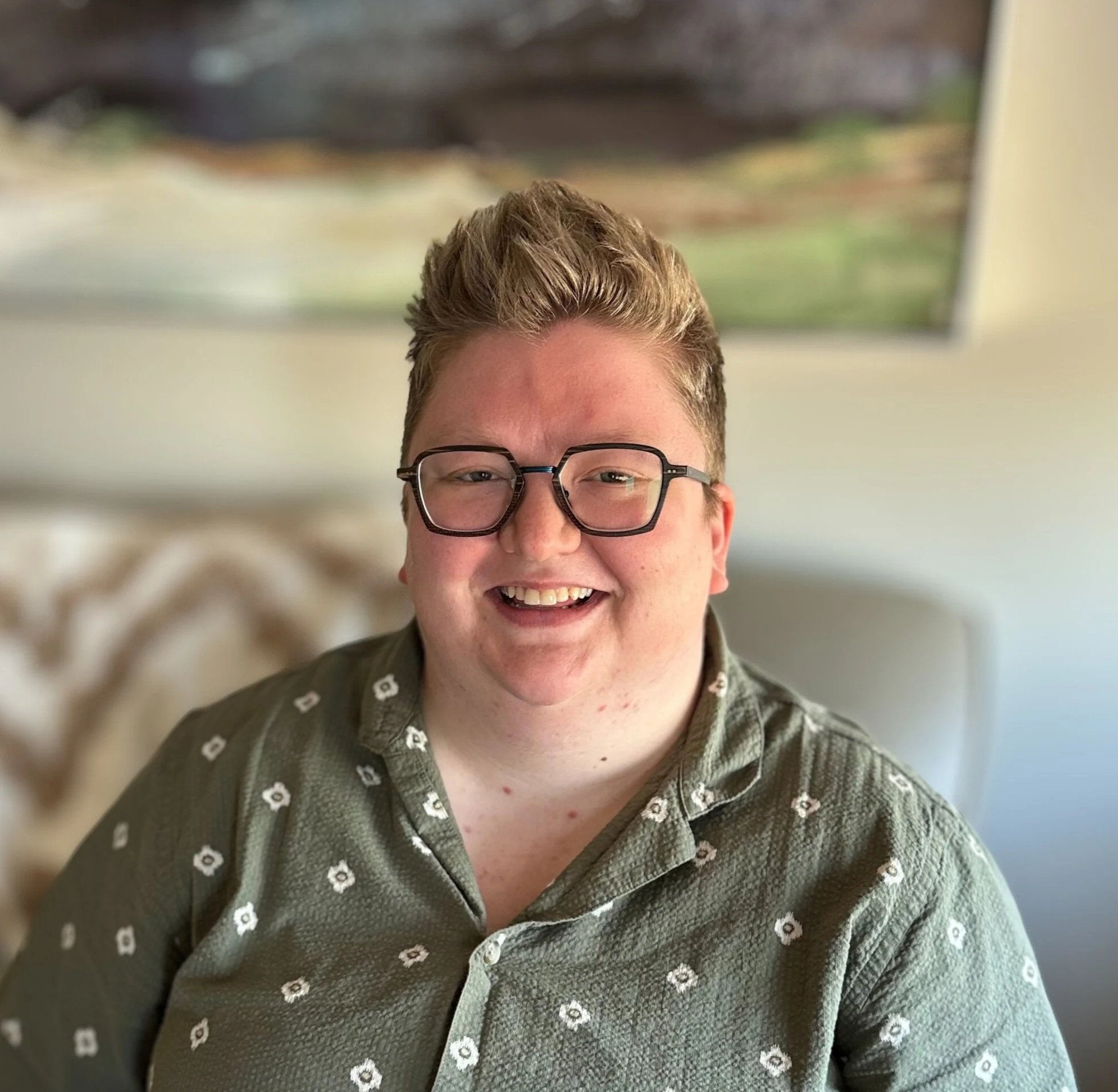 A smiling young man with glasses and short, styled hair, wearing a patterned green shirt, sitting on a beige couch in a well-lit room with a blurred landscape painting on the wall behind him.