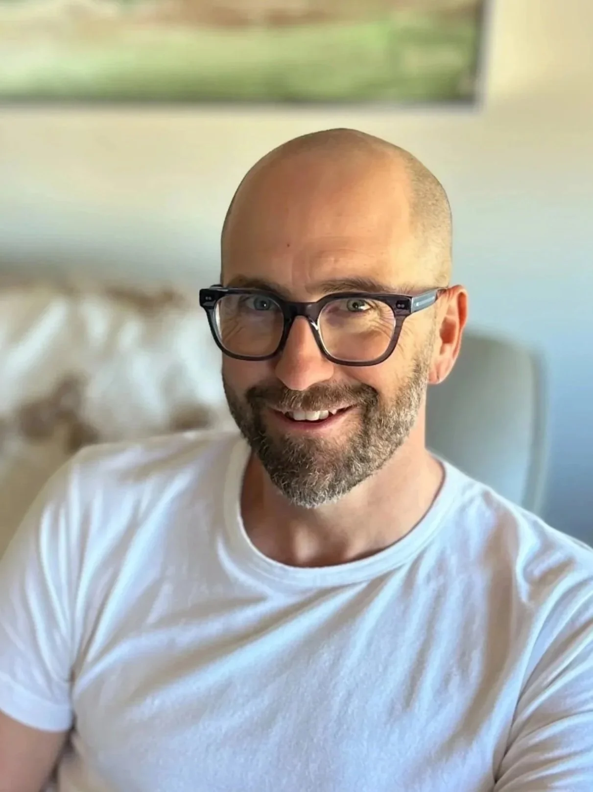 A smiling bald man with glasses and a beard, wearing a white t-shirt, sitting in a room with a blurred background and natural light.