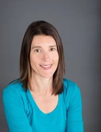 Portrait of a woman with shoulder-length dark hair, smiling, wearing a bright blue top against a gray background.