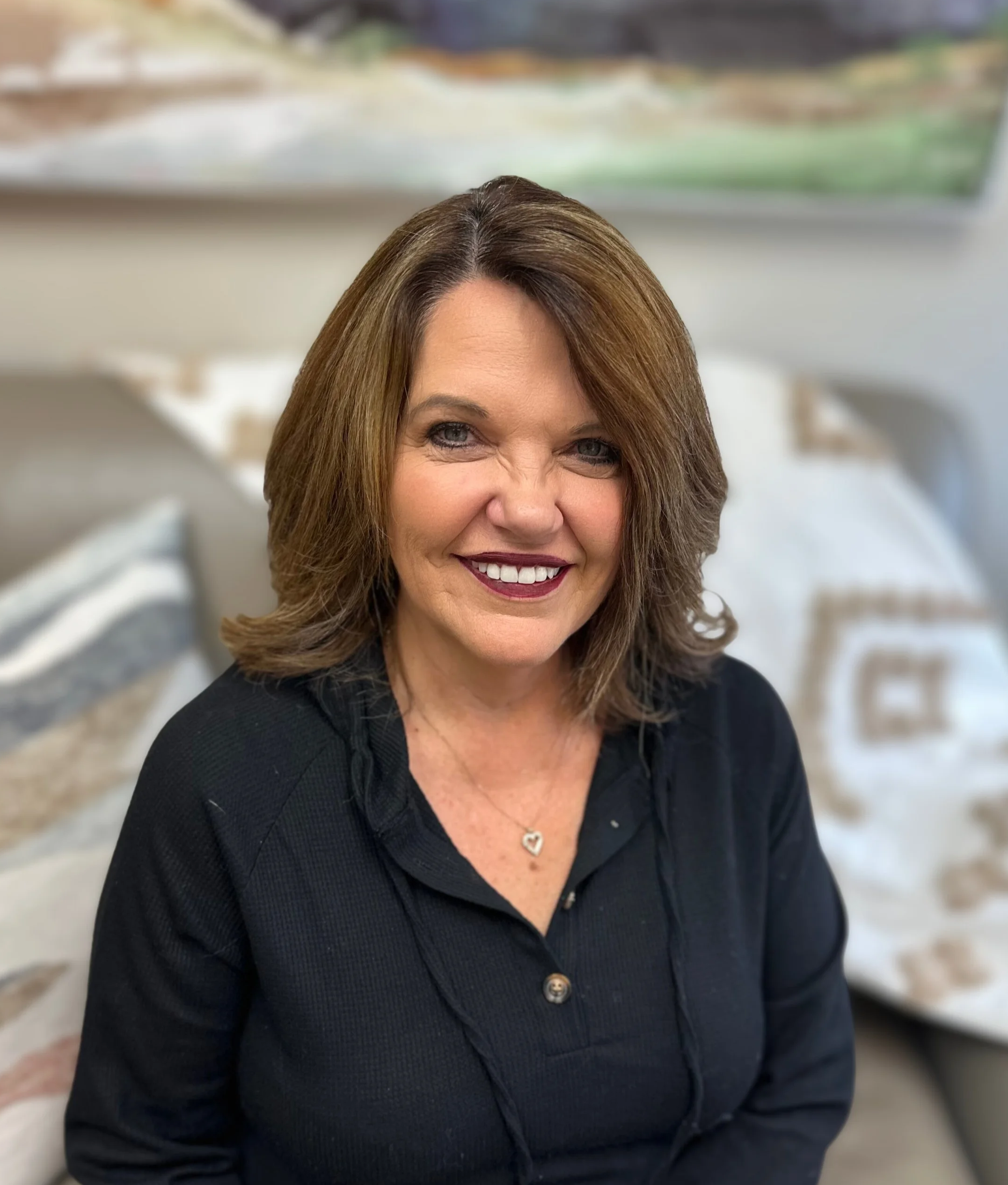 A woman with shoulder-length brown hair, smiling broadly, wearing a black top and a silver heart pendant necklace, sitting on a sofa with decorative pillows in the background.
