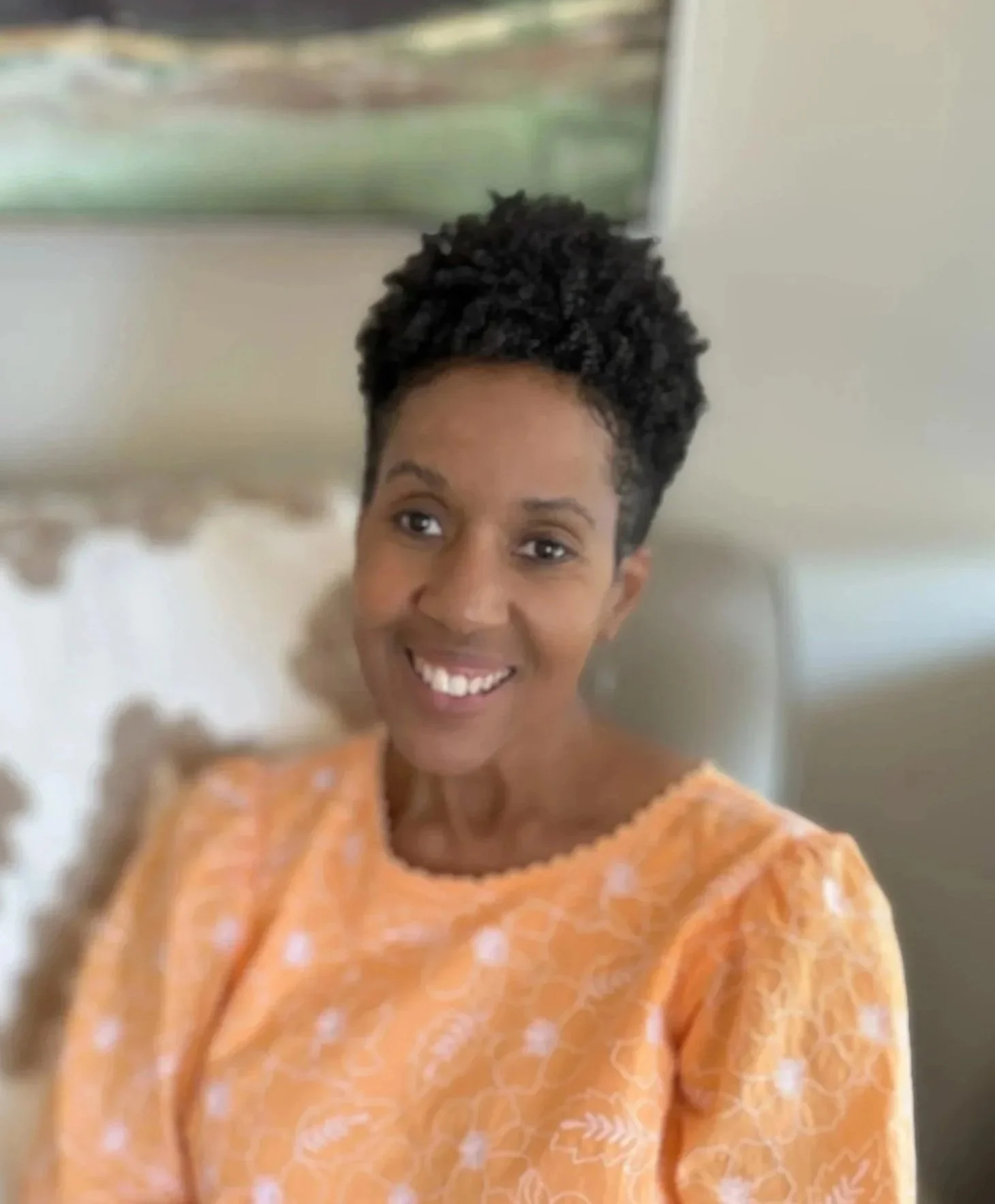 A woman with dark skin and short curly hair smiling while sitting on a bed in a room with a lamp and a window in the background.