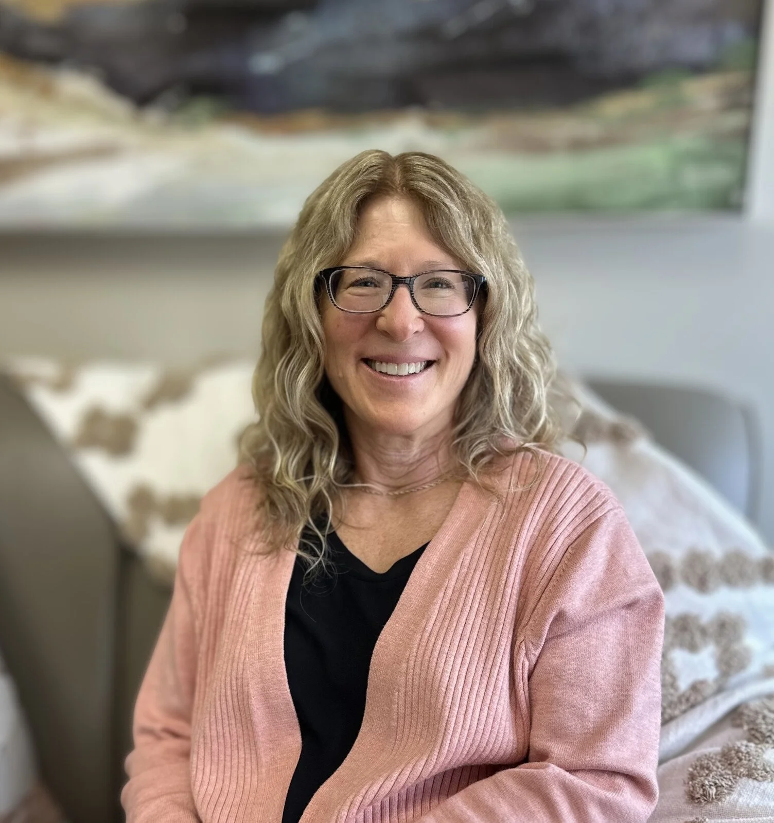 A woman with curly blonde hair, wearing glasses, a black top, and a pink cardigan, smiling and sitting on a sofa with patterned pillows in a living room.