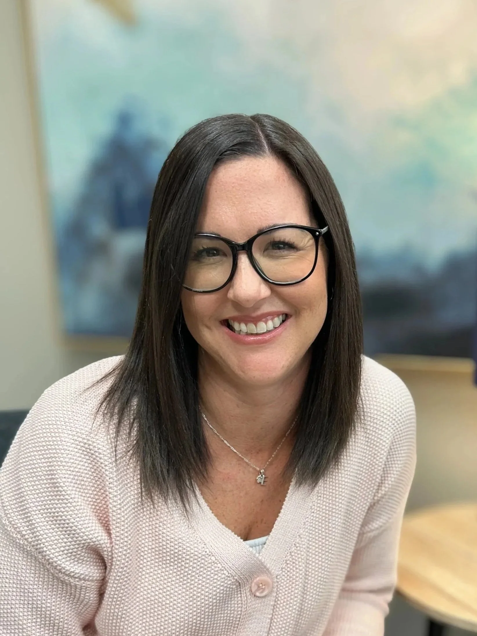 A smiling woman with dark hair and glasses, wearing a light pink sweater, sitting indoors with a blurred abstract painting in the background.