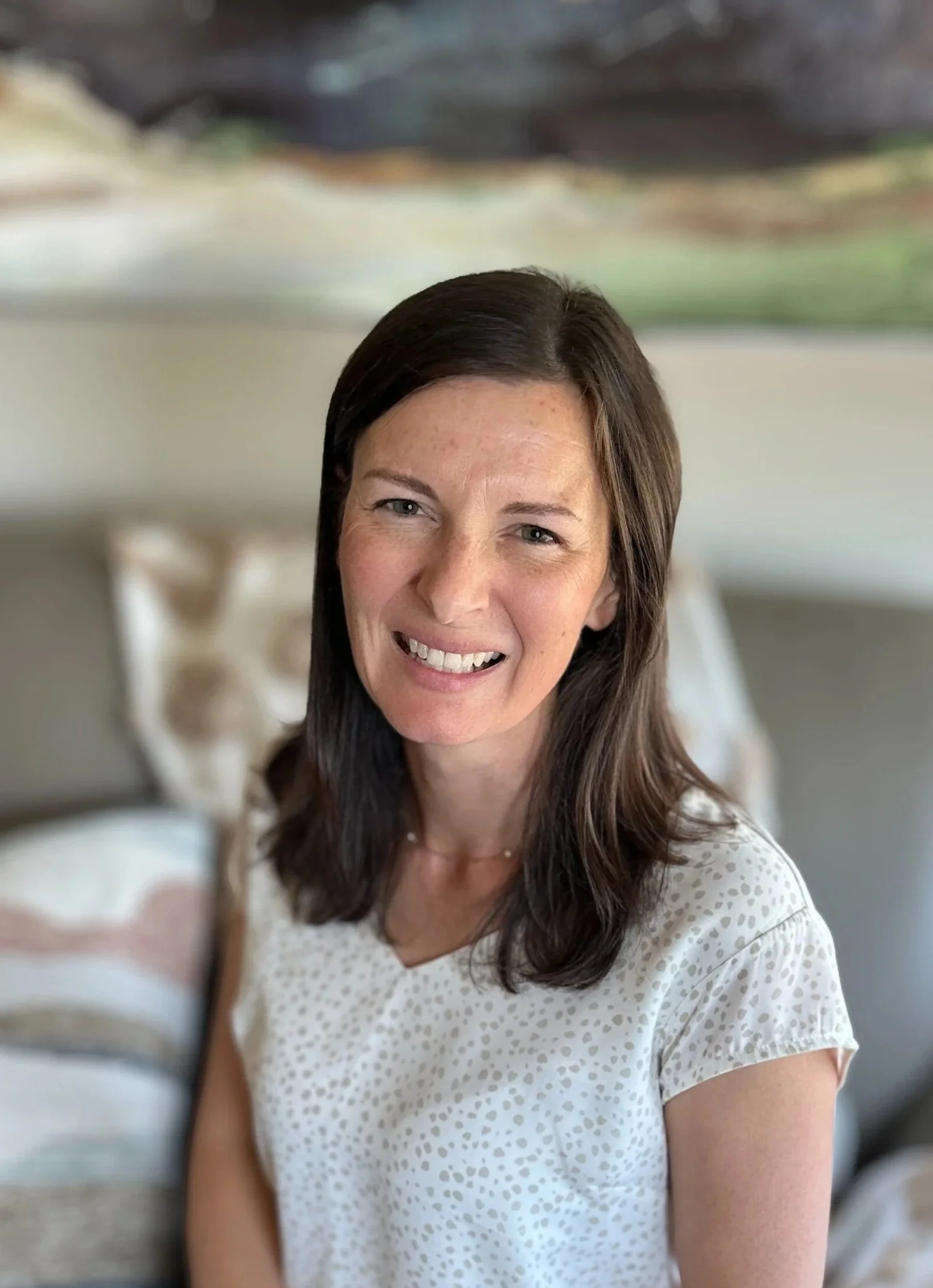 A woman with shoulder-length dark brown hair smiling, sitting indoors with a blurred abstract artwork and a sofa in the background, wearing a white dotted blouse.