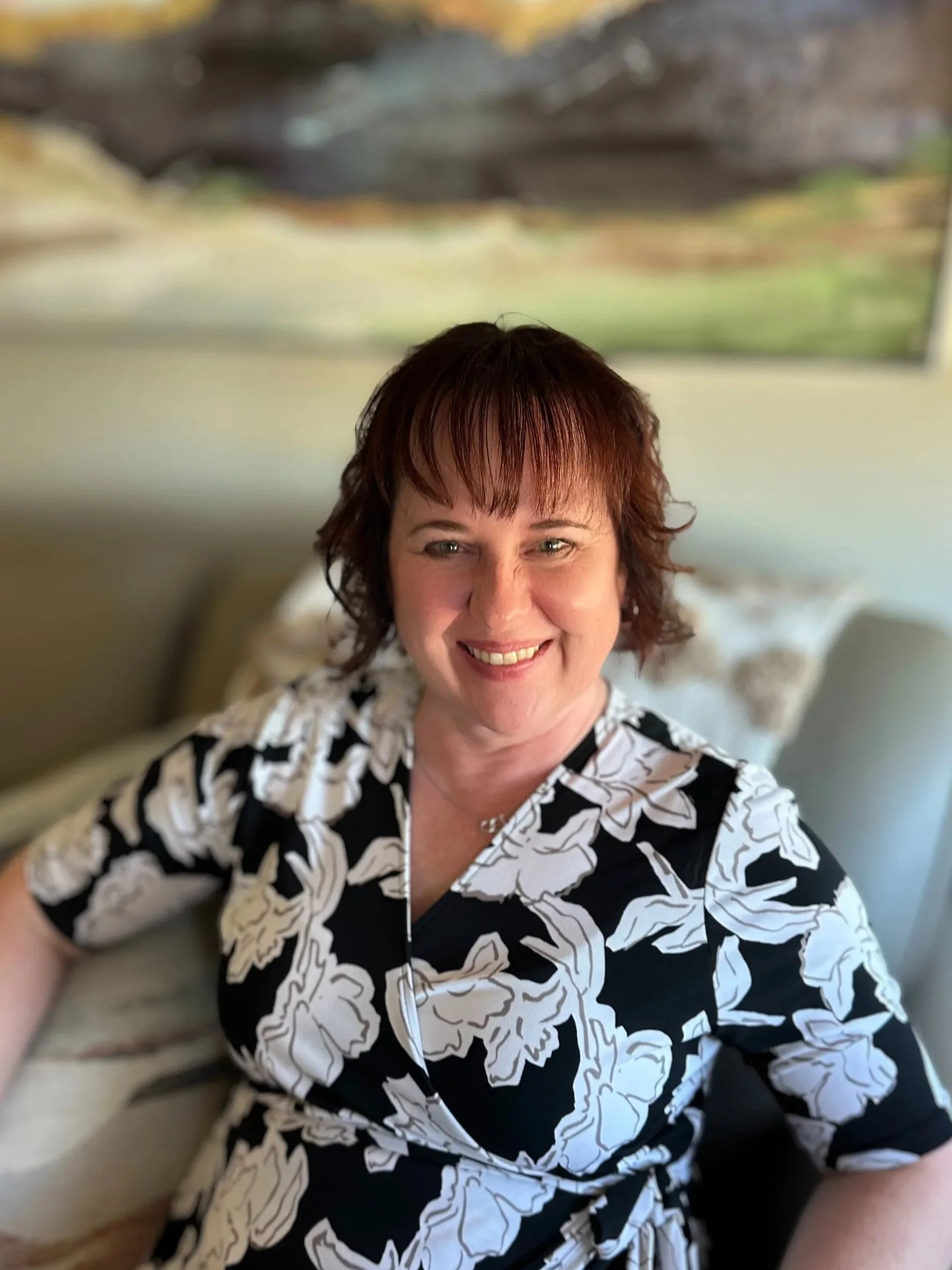 A woman with short reddish-brown hair smiling, wearing a black and white floral dress, sitting on a couch with a framed picture or painting of a nature scene behind her.